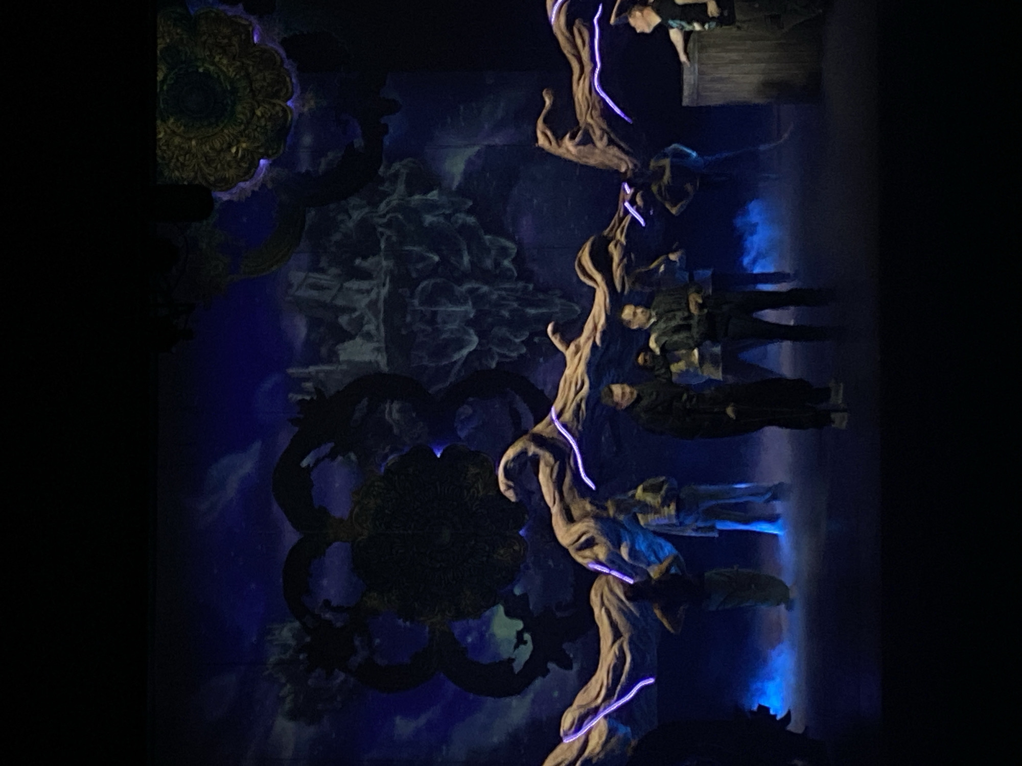 The image depicts a scene from a performance, possibly a theatrical production, in Rusanovo, Russia. The backdrop features intricate floral patterns and stylized rock formations in shades of blue and black. A large, artificial tree with illuminated purple accents is positioned prominently. Several figures are visible, appearing to be performers in costume. Some stand near the tree, while others are suspended or positioned higher in the scene, creating a multi-layered visual effect. A structure resembling a small building or platform is partially visible in the upper right corner. The overall lighting is theatrical, emphasizing the scene's dramatic and fantastical qualities.