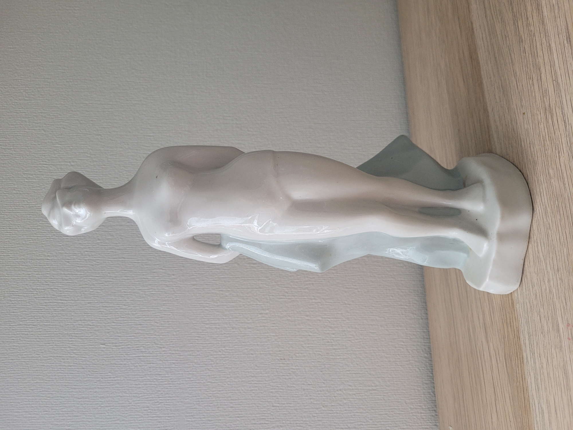 A white porcelain figurine of a nude woman stands on a light wood surface against a textured pale gray wall. The figurine is smooth and stylized, with her hair pulled up. She is draped with a light blue fabric that covers part of her body and extends down to the base. The figurine's base is irregular and white, also appearing to be made of porcelain. The background wall has a subtle vertical texture. The scene is indoors, with no indication of a specific city or location.