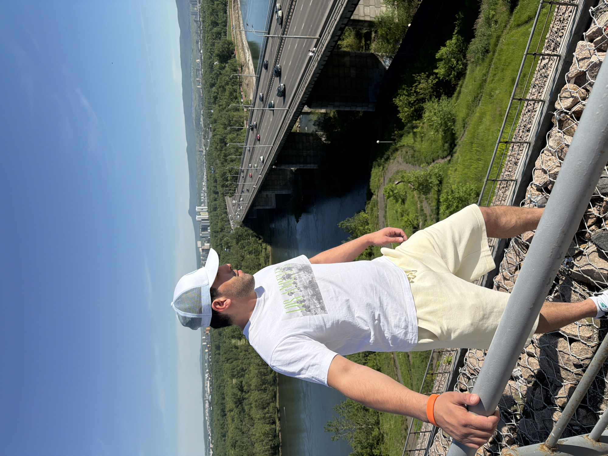 The image shows a male figure standing on an elevated observation point overlooking the Yenisei River and the Nikolaevsky Bridge in Krasnoyarsk, Russia. The man, viewed from the mid-thigh up and facing towards the right side of the frame, has light skin, dark hair, and visible facial stubble. He wears a white baseball cap with a light blue mesh back, a white short-sleeved t-shirt featuring a "LOS ANGELES" graphic with palm trees in black and green, and light yellow shorts. An orange wristband is present on his left wrist as he grips a grey metal railing in the foreground. Beneath the railing, a gabion wall filled with rocks is visible, supporting a steep, vegetated slope that descends towards the river.
The wide, dark green Yenisei River flows from left to right across the mid-ground. Spanning the river is a multi-lane concrete bridge, identified as the Nikolaevsky Bridge, with multiple support piers. The bridge carries numerous cars in both directions and includes a pedestrian walkway. Beyond the bridge and the river, the urban landscape of Krasnoyarsk is visible, characterized by numerous multi-story buildings interspersed with green areas. Distant hills or low mountains form the horizon line. The sky above is a clear, bright blue with a few faint wisps of clouds. The scene is illuminated by strong daylight.