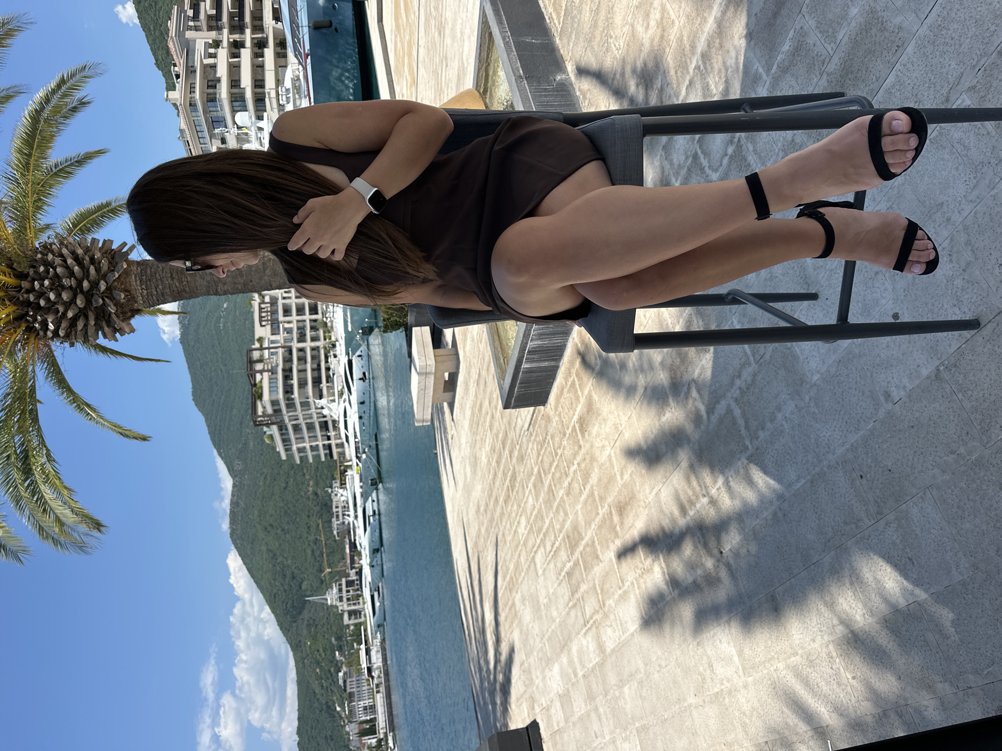 A woman with long, dark hair sits facing right on a tall, grey bar stool. She is wearing a dark brown, sleeveless dress and black, ankle-strap sandals. She is wearing sunglasses and has her right hand touching her hair. The woman is positioned in the foreground, and behind her, a body of water is visible, lined with white yachts and buildings. Mountains rise in the background, covered in green foliage. A palm tree is present to the left of the woman, casting a shadow on the paved ground. The scene appears to be a waterfront promenade in Donja Lastva, Montenegro, likely a marina or harbor area given the presence of yachts. The lighting suggests it is a sunny day.