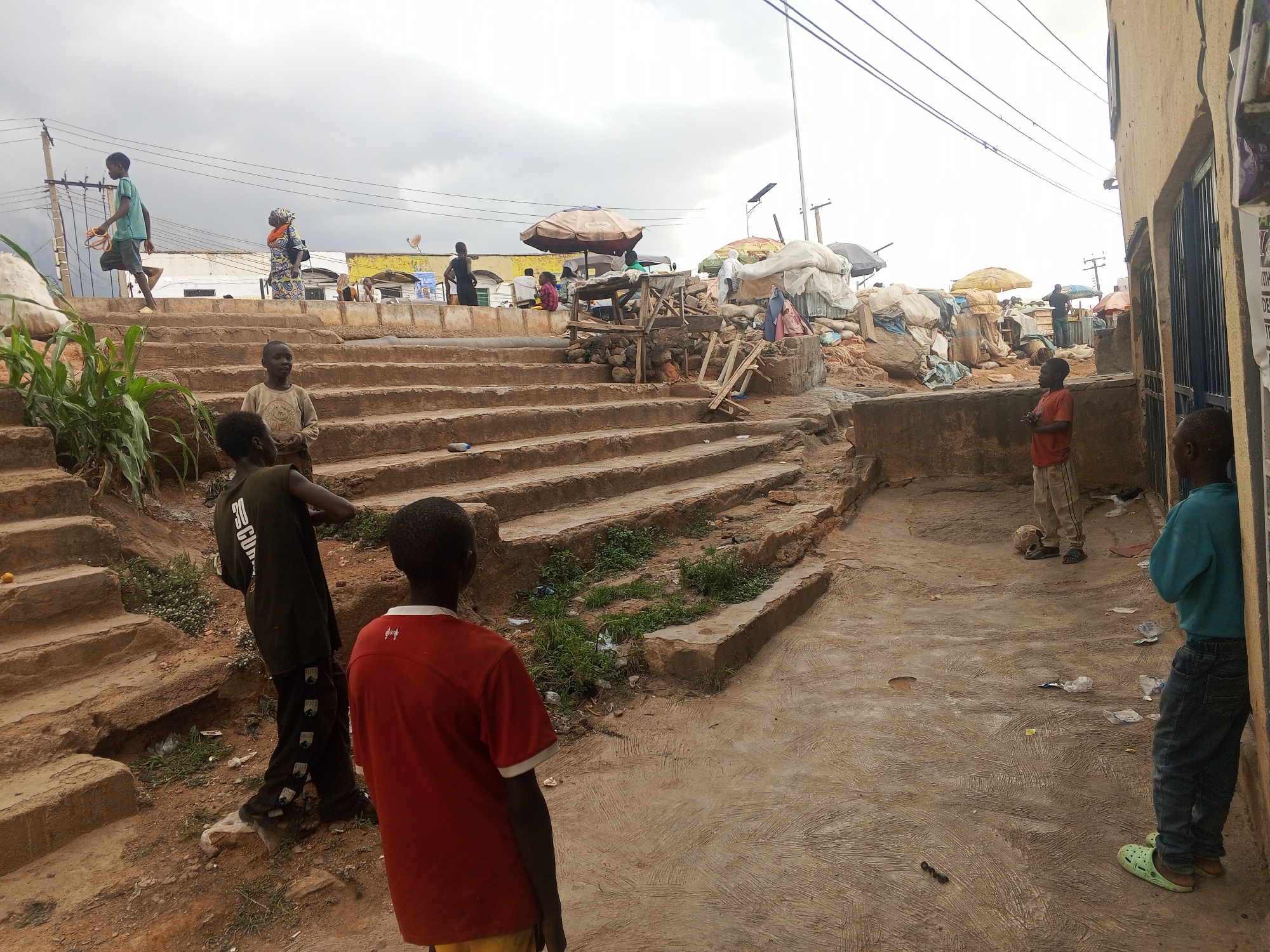 The image depicts a scene in Gwong, Nigeria. Several children are visible: two stand near a concrete structure to the right, one in a red shirt, another in a dark shirt is to the left, another stands on the steps in the background, and one walks up the steps. Behind the steps, people are gathered near market stalls covered by umbrellas. The ground is uneven and strewn with litter. Overhead power lines are visible. The sky is overcast.