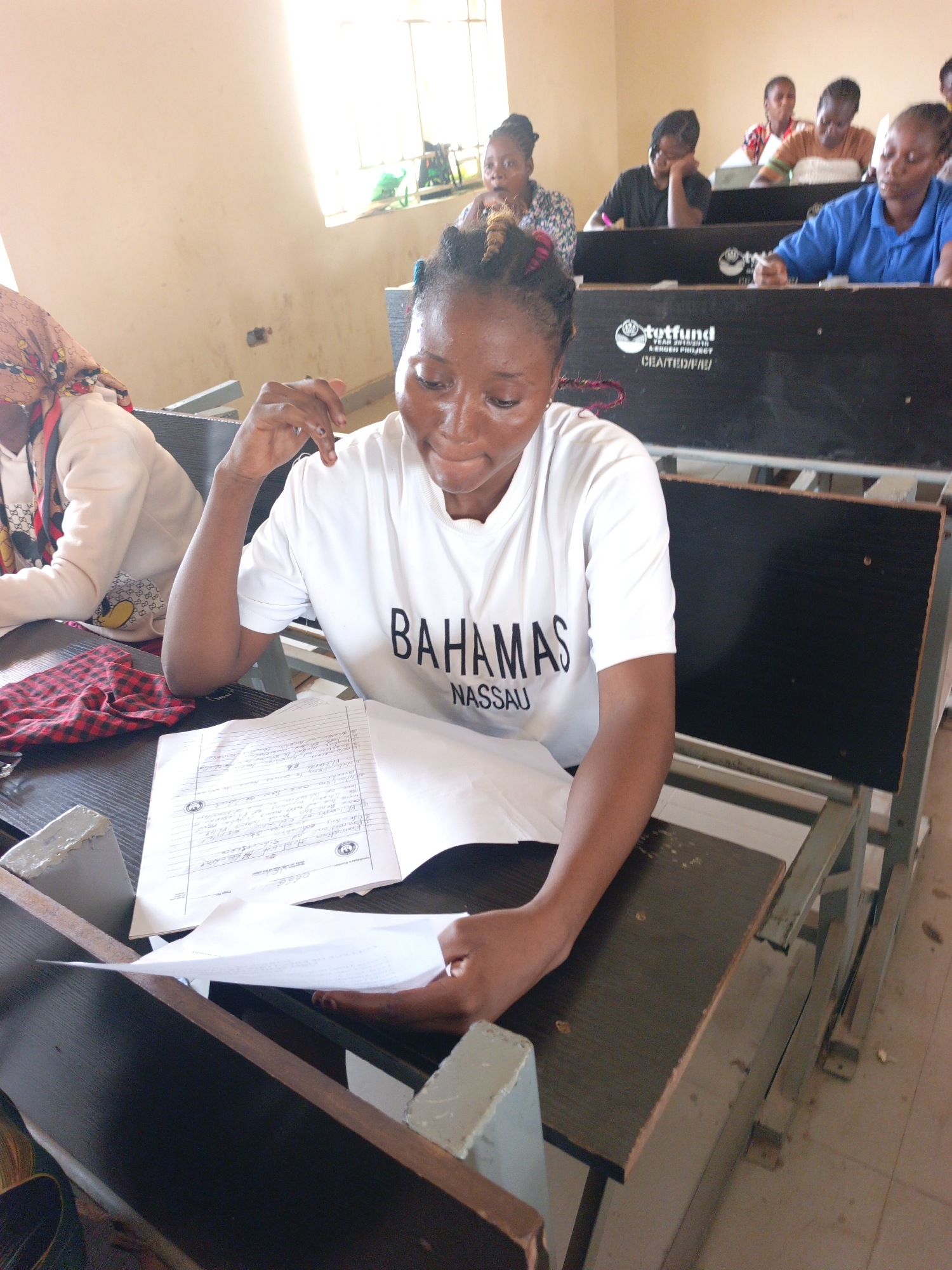 The image depicts a classroom setting, possibly in Akwanga, Nigeria, based on the student population and the presence of "totfund" branded desks, a developmental project. The foreground shows a woman in a white t-shirt with "BAHAMAS NASSAU" printed on it, examining handwritten papers at her desk. She is positioned in the front row of a tiered classroom. Several other students are present in the background, some engaged in similar activities. The desks are wooden and appear well-used. Natural light enters the room from a window on the upper left.