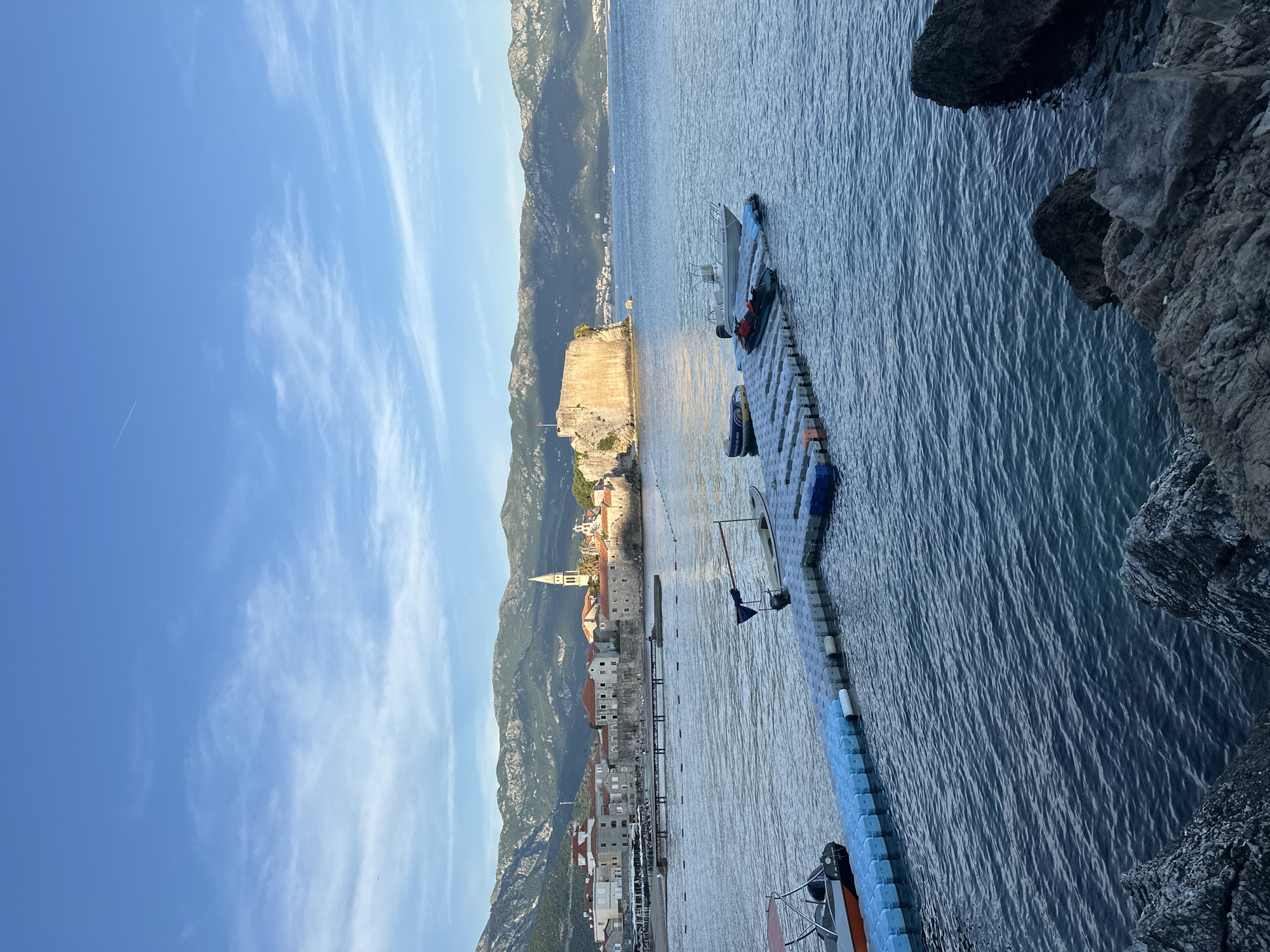 The image captures a waterfront view of Budva, Montenegro, under a partly cloudy, bright blue sky. In the background, a mountainous range stretches, providing a scenic backdrop to the urban landscape. In the middle ground is a medieval walled city with buildings of red-tiled roofs and a prominent church spire. An ancient stone fortress is situated on the edge of the water. The foreground contains a blue plastic modular dock or platform extending into the water. The water itself displays gentle ripples, reflecting the ambient light. The scene is captured from an elevated position, likely a rocky cliffside.