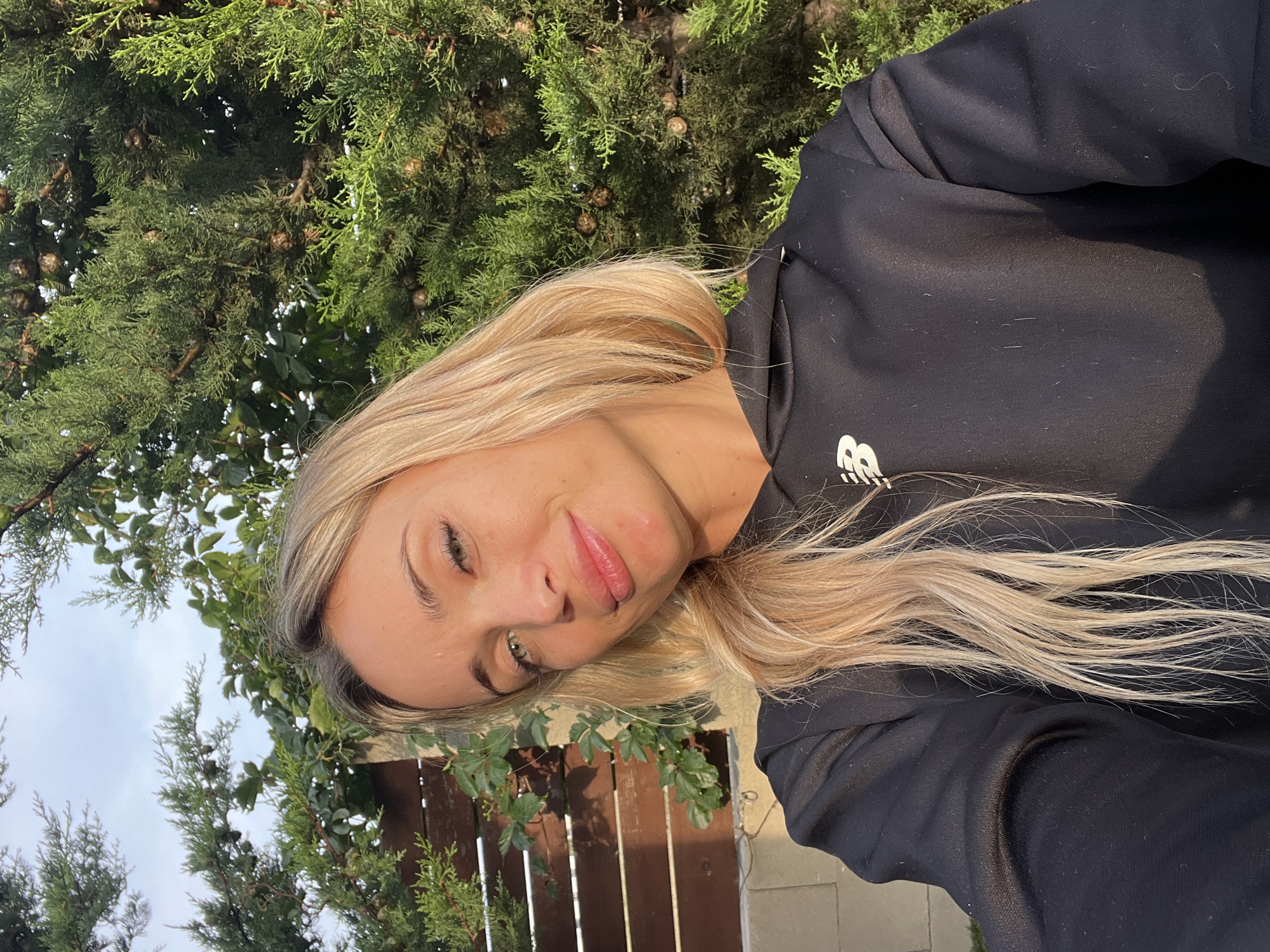 A woman with blonde hair smiles at the camera, taking a selfie outdoors. She is wearing a black hoodie with a white logo. Behind her, there is a wooden fence with ivy growing on it, and a large green tree with cones. The lighting suggests it is either early morning or late afternoon. The location is Tsodoreti, Georgia.