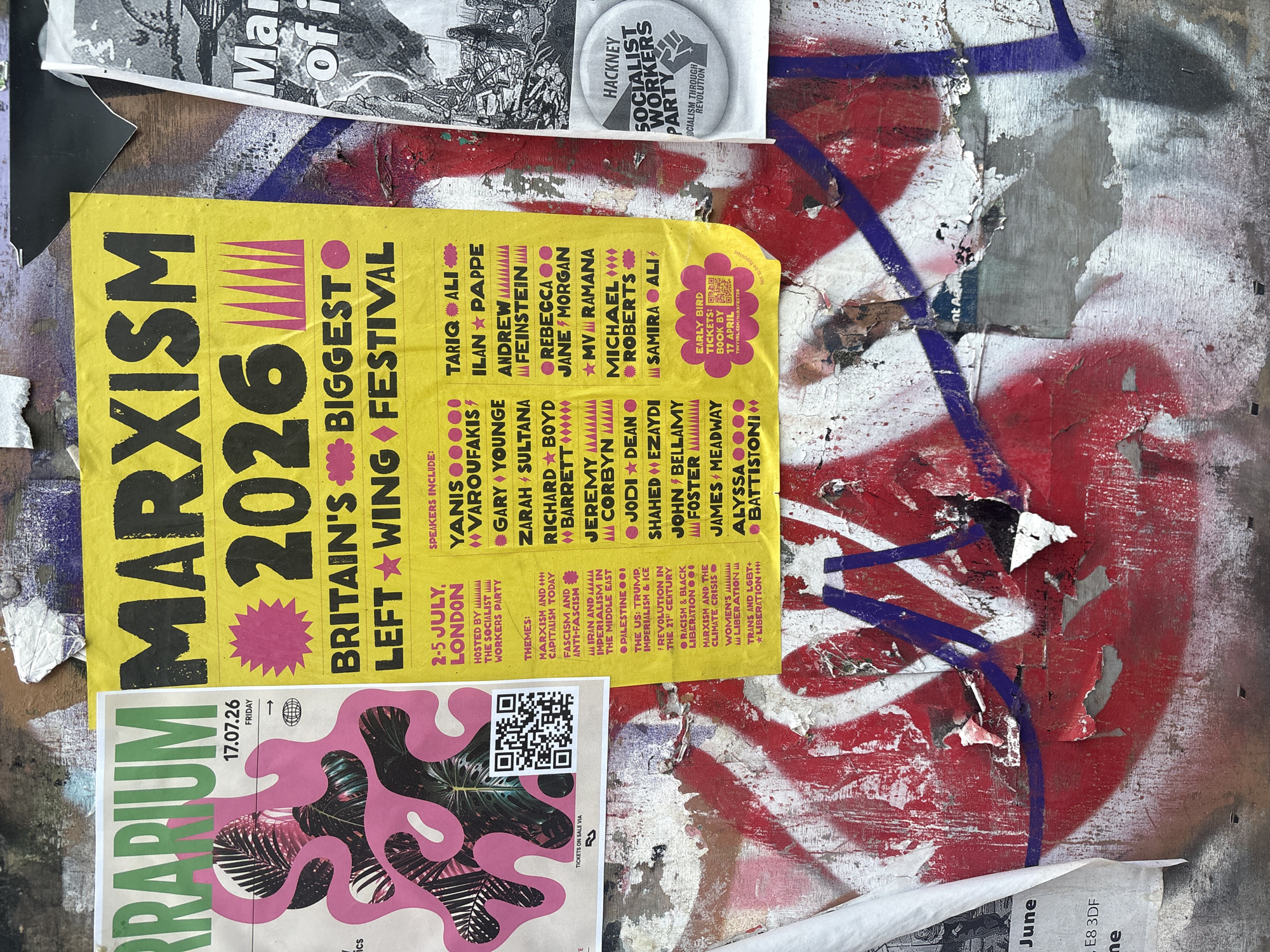 The image shows a close-up of two posters plastered on a weathered, graffitied wall. The poster on the left, with a pink and green abstract design, reads "RRARIUM" and appears to be promoting an event on "17.07.26 FRIDAY." A QR code is visible. The larger poster on the right is bright yellow and displays bold black text: "MARXISM 2026 BRITAIN'S BIGGEST LEFT WING FESTIVAL." It lists event details including "2-5 JULY, LONDON" and names various speakers and themes such as "FASCISM AND ANTI-FASCISM" and "THE US: TRUMP, IMPERIALISM & ICE." A smaller circular badge with "HACKNEY SOCIALIST WORKERS PARTY SOCIALISM THROUGH REVOLUTION" is attached to the right side of the yellow poster. The wall itself is covered in layers of peeling paint, graffiti in red and purple, and other torn paper fragments, suggesting an urban outdoor setting in London.