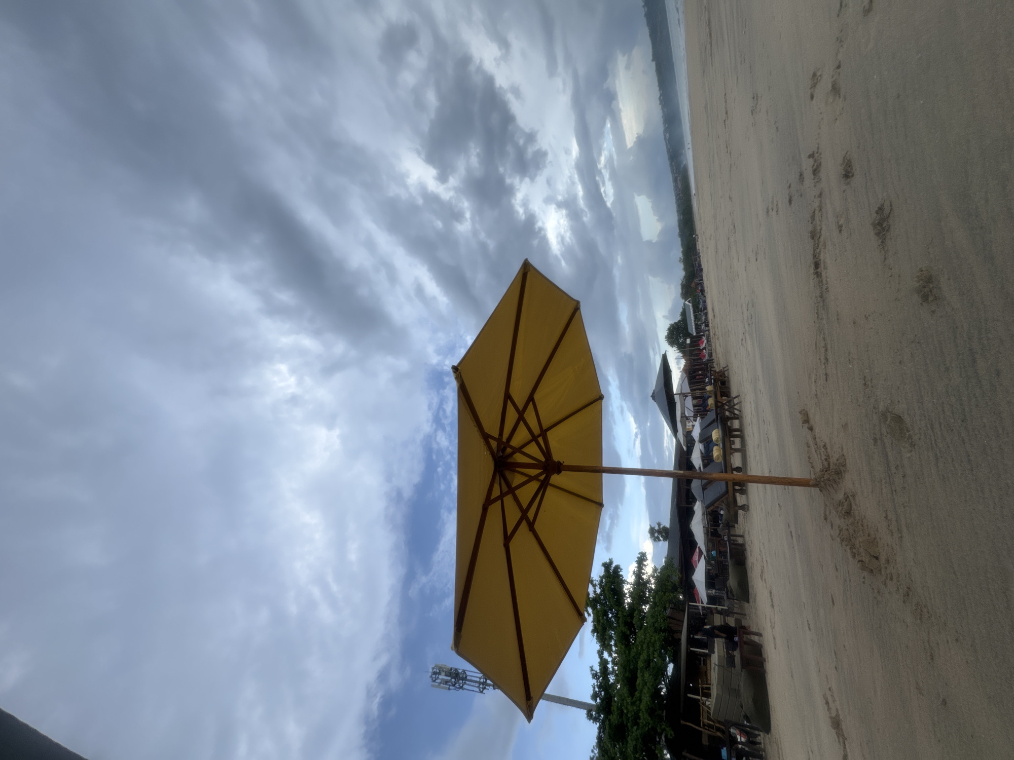 The image displays a serene beach scene, likely situated in Kuta Selatan, Indonesia, under a dynamic sky. The foreground is dominated by a large, open yellow beach umbrella, its wooden support structure visible underneath, positioned slightly to the left of the frame's center. To the left of the umbrella, a tall communication tower rises against the sky.
The right side of the image features expansive sandy beach, marked with numerous footprints that recede into the distance. Along the left edge of the beach, several smaller beach umbrellas, along with chairs and tables, are arranged, indicating beachside establishments. Green trees and foliage are visible amongst these structures. The distant ocean and coastline appear as a hazy, narrow band at the far right edge, blending with the horizon.
The sky is a prominent feature, showcasing a mix of dramatic grey clouds and intermittent patches of clear blue, suggesting an overcast day or perhaps late afternoon light. No individuals are distinctly visible, and the scene appears calm and quiet. There is no visible text within the image. The overall impression is one of a tranquil coastal setting.
