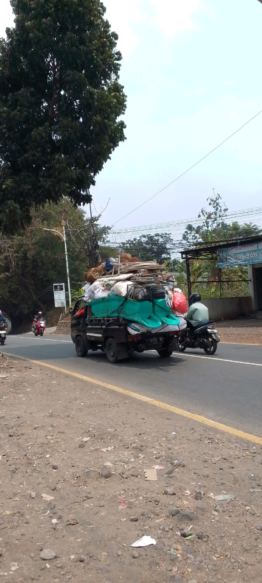 An outdoor, eye-level, medium shot captures a dark-colored pickup truck heavily laden with goods. The truck is driving on a paved road, which appears to be in a rural or semi-urban area, indicated by the presence of trees, vegetation, and a building in the background. The load on the truck is diverse, including what appears to be green tarpaulin, bundles of wood, sacks, and what look like large, curved plastic items. One red sack is prominently visible. In the foreground, the road's shoulder is rough and dirt-covered with scattered debris.
To the right of the pickup truck, a person wearing a light-colored shirt and a helmet is riding a black motorcycle, moving in the same direction. Further back on the left side of the road, two more motorcycles are visible, also heading in the same direction. One motorcycle has a passenger, and both appear to be passing the truck.
The background includes trees and a building with signage. The building has a dark awning and text on its facade, partially obscured. A utility pole with wires is also visible. The sky is overcast and pale. The scene is consistent with a location along a road in Cilaku, Indonesia, likely a transportation route for goods. The license plate of the pickup truck appears to read "9172 WP".