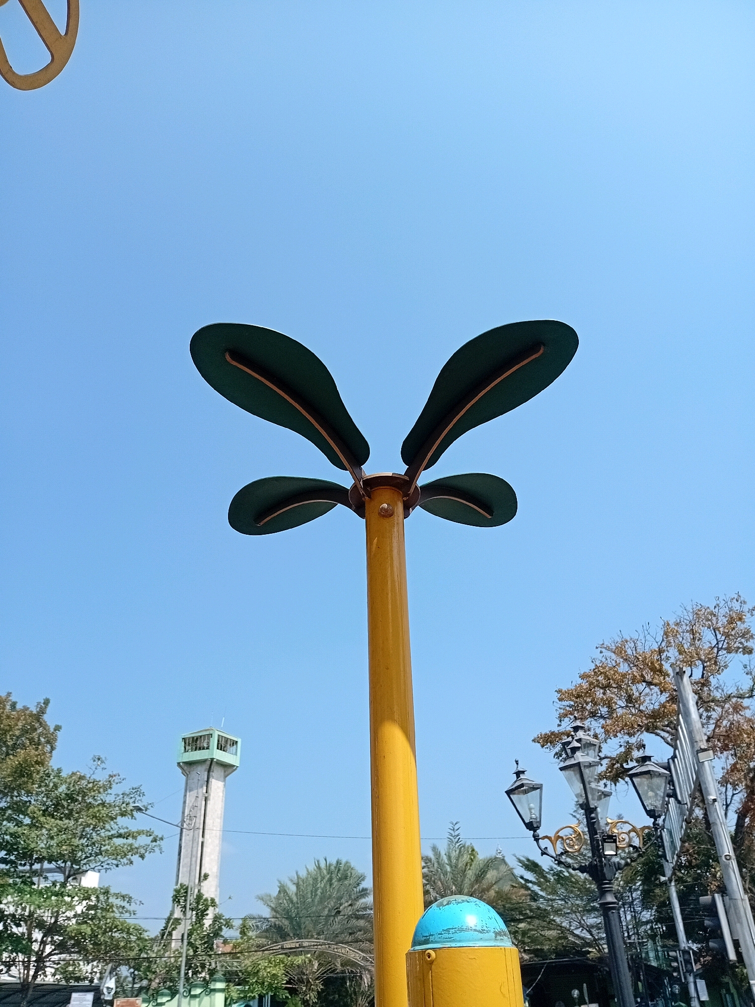 A low-angle, full shot captures a bright, clear blue sky above a park-like setting in Purworejo, Indonesia. In the foreground, a yellow pole stands tall, adorned with four large, dark green leaf-shaped decorations at its apex. To the left of the main pole, a segment of a large, circular, light brown object is visible against the sky. Further back, a tall, slender concrete tower with a light green, cage-like structure at its summit pierces the horizon, flanked by lush green trees and palm trees. To the right of the tower, an ornate black lamppost with three glass-shaded lamps stands amongst dry, brown-leafed trees. A bright blue, rounded object sits atop a portion of the yellow pole in the lower right corner of the image. The overall scene is tranquil and sunny, suggesting a pleasant day in an outdoor public space.