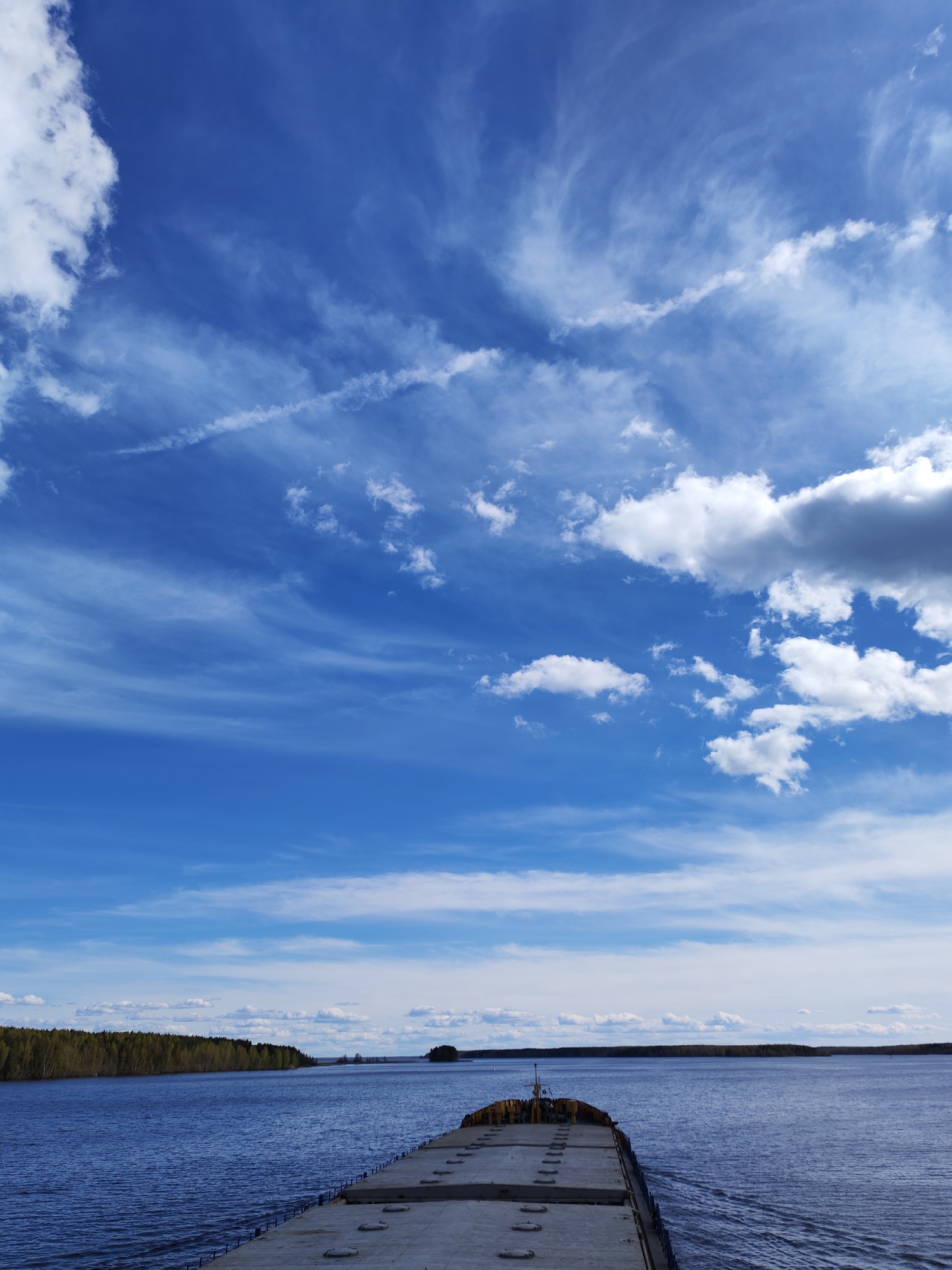 The image depicts a wide-angle view from the stern of a large barge moving across a body of water, likely a river or large lake. The sky dominates the upper portion of the frame, showcasing a vibrant blue hue interspersed with various cloud formations including wispy cirrus and cumulus clouds. A distinct contrail is visible as well. The barge itself is made of concrete. The body of water reflects the sky's color and the surrounding environment. On the left bank of the water body, a dense green forest line runs parallel to the shore. A small island is visible in the distance.
While the image contains elements typical of a waterfront landscape, there is no definitive visual information present in the image to confirm a specific location such as Topornya, Russia.