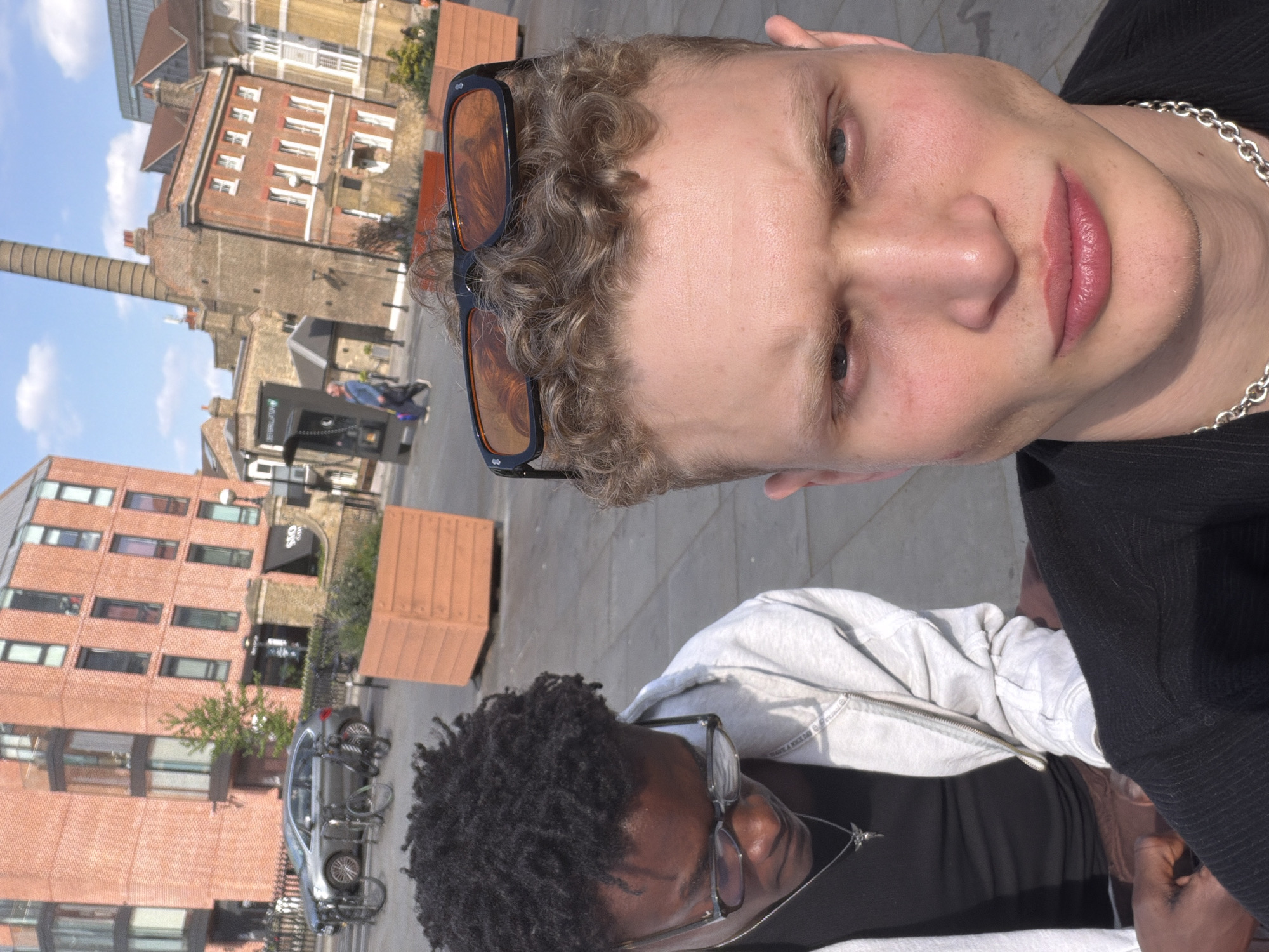 The image, rotated 90 degrees counter-clockwise, presents a close-up, high-angle shot of two individuals with an urban London backdrop on a sunny day.
The primary subject, occupying the foreground, is a light-skinned person with curly, light blonde hair. They wear dark-framed sunglasses pushed up onto their head and a silver chain necklace. Their facial features include a prominent nose, full, pink-tinted lips, and light-colored eyes (likely blue or grey). Their expression is neutral, looking directly towards the viewer. They are wearing a dark, possibly black, ribbed top or jacket.
Behind and below the main subject, partially obscured, is a second person with darker skin and dark, textured curly hair. This individual wears clear-framed glasses and a light grey hooded sweatshirt over a dark t-shirt, with a silver necklace visible. Faint text, possibly "HAVE A NICE DAY," is subtly visible on the collar area of their hoodie.
The background reveals a bustling urban environment typical of London. Predominantly brick buildings in shades of red, brown, and tan line the streets. A tall, slender brick chimney stands out against the bright blue sky. A silver car with bicycles mounted on its rear is parked on the street in front of one of the buildings. One building features a sign with the letters "OEO" above an arched entrance. The bright sunlight casts distinct shadows, indicating a clear, sunny day. Wooden planters or benches are also visible in the street scene. The overall setting suggests a lively city area, potentially with a mix of residential, commercial, and historical industrial architecture.