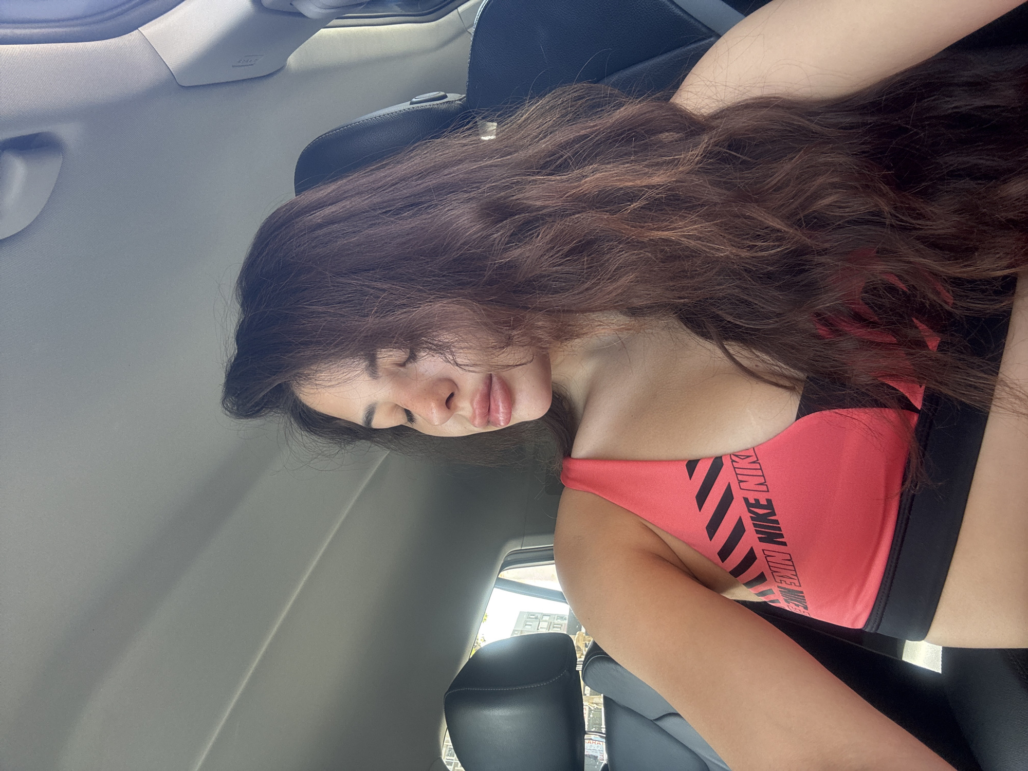 A medium shot, indoor photograph shows a young woman with long, wavy, dark brown hair seated in the passenger seat of a car. She is wearing a pink and black sports bra with a Nike logo. Her eyes are closed, and her head is tilted back slightly. The interior of the car is visible, including the headrest of the driver's seat and the car's headliner. The window on the left side of the frame shows a blurred view of a building and some trees, suggesting an urban environment. The lighting indicates it is daytime.