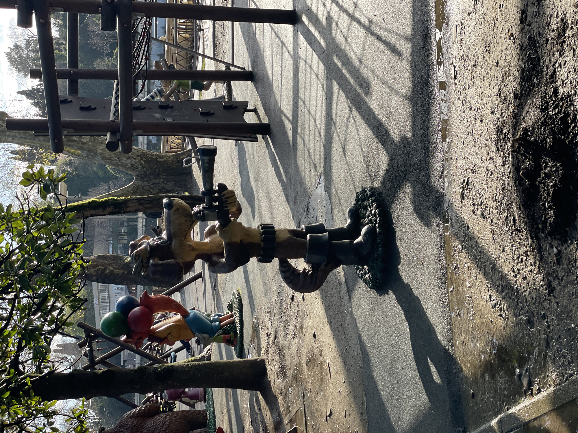 The image depicts a playground in Sochi, Russia. There are several wooden structures for children to climb on and play with. A statue of a character with a camera is visible, along with other painted figures holding balloons. The ground appears to be a combination of gravel and dirt, with some wet areas. The lighting suggests a sunny day, and there are shadows cast by the structures and figures. Trees and buildings are visible in the background.