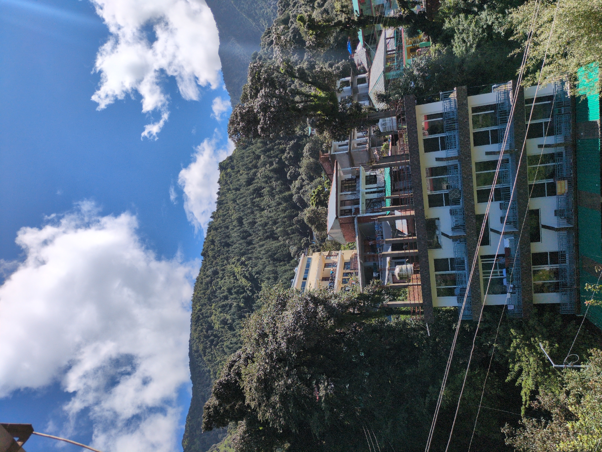 The image shows a multi-story hotel situated on a hillside in Bhagsu, India. The hotel has a white facade with balconies on each floor, featuring large glass windows and railings. Several rooms appear to have furniture visible on the balconies, including tables and chairs, and some laundry is also hanging to dry. Power lines crisscross the foreground, partially obscuring the hotel and the surrounding lush green trees and dense forest covering the mountainous terrain behind. The sky is a bright blue with scattered white clouds, indicating a clear and sunny day. Several other buildings, also on the hillside, are visible in the background, some with colorful rooftops. The architecture suggests a warm climate and a tourist-oriented location.