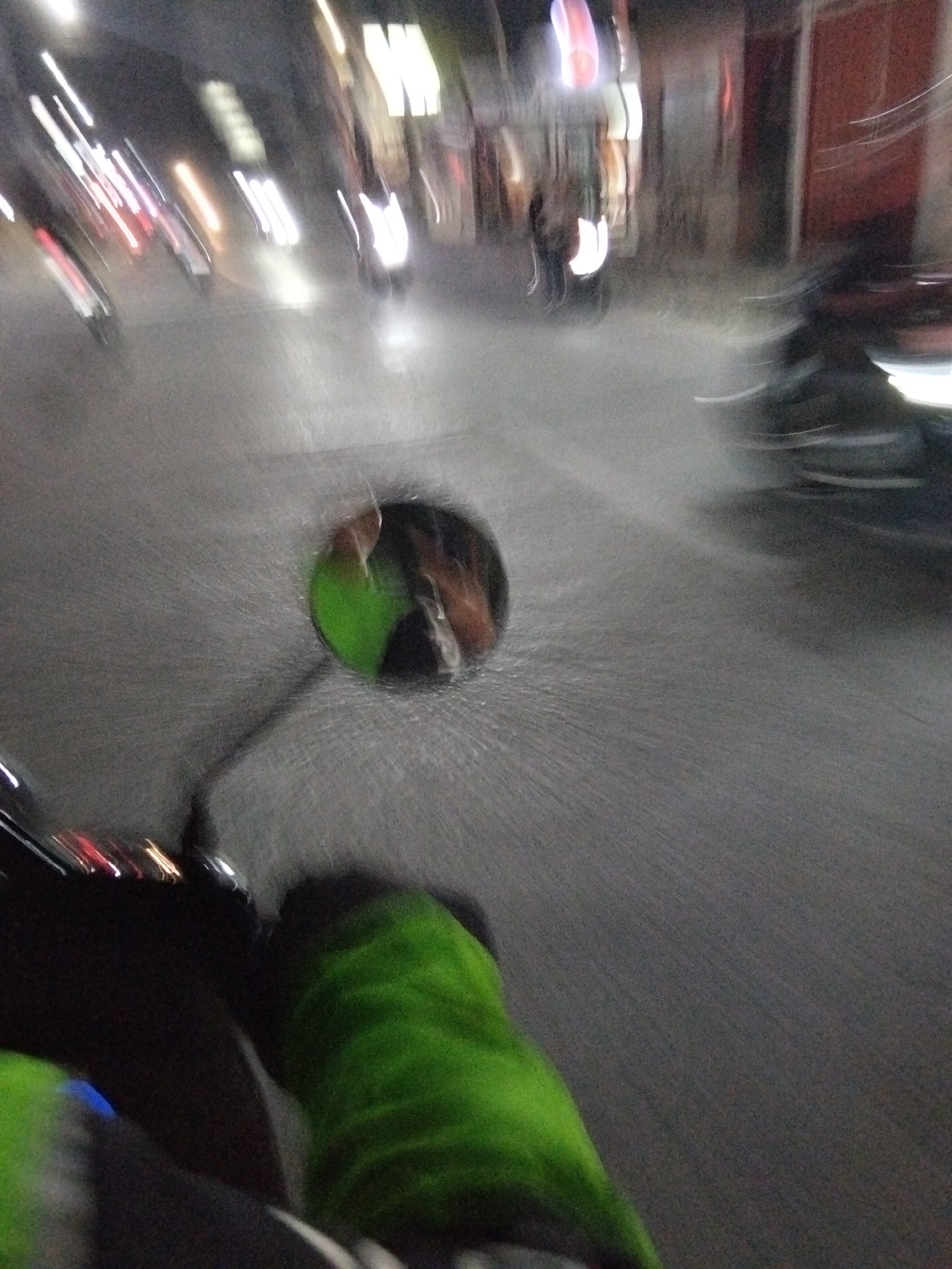 The image is a motion-blurred photograph taken at night on a street in South Tangerang, Indonesia. The perspective suggests the photo was taken from a moving vehicle, likely a motorcycle, looking forward.
In the foreground, the handlebars of the vehicle are visible on the left, with a bright green fabric covering a portion of them. Part of the rider's leg, also covered in bright green fabric, is visible at the bottom right of the frame.
The central focus of the image is a circular rearview mirror mounted on the vehicle. Reflected in the mirror is a blurry image of the rider's head and upper body. A green object, possibly part of the rider's clothing or an accessory, is also visible in the reflection.
The background depicts a city street scene at night, characterized by streaking lights from other vehicles and buildings. Several blurred vehicles, appearing as streaks of light, are moving on the road. The road surface is visible, appearing to be asphalt. Buildings lining the street are dimly lit, with some illuminated signs. The overall impression is one of movement and the dynamic environment of a nocturnal urban setting in South Tangerang.