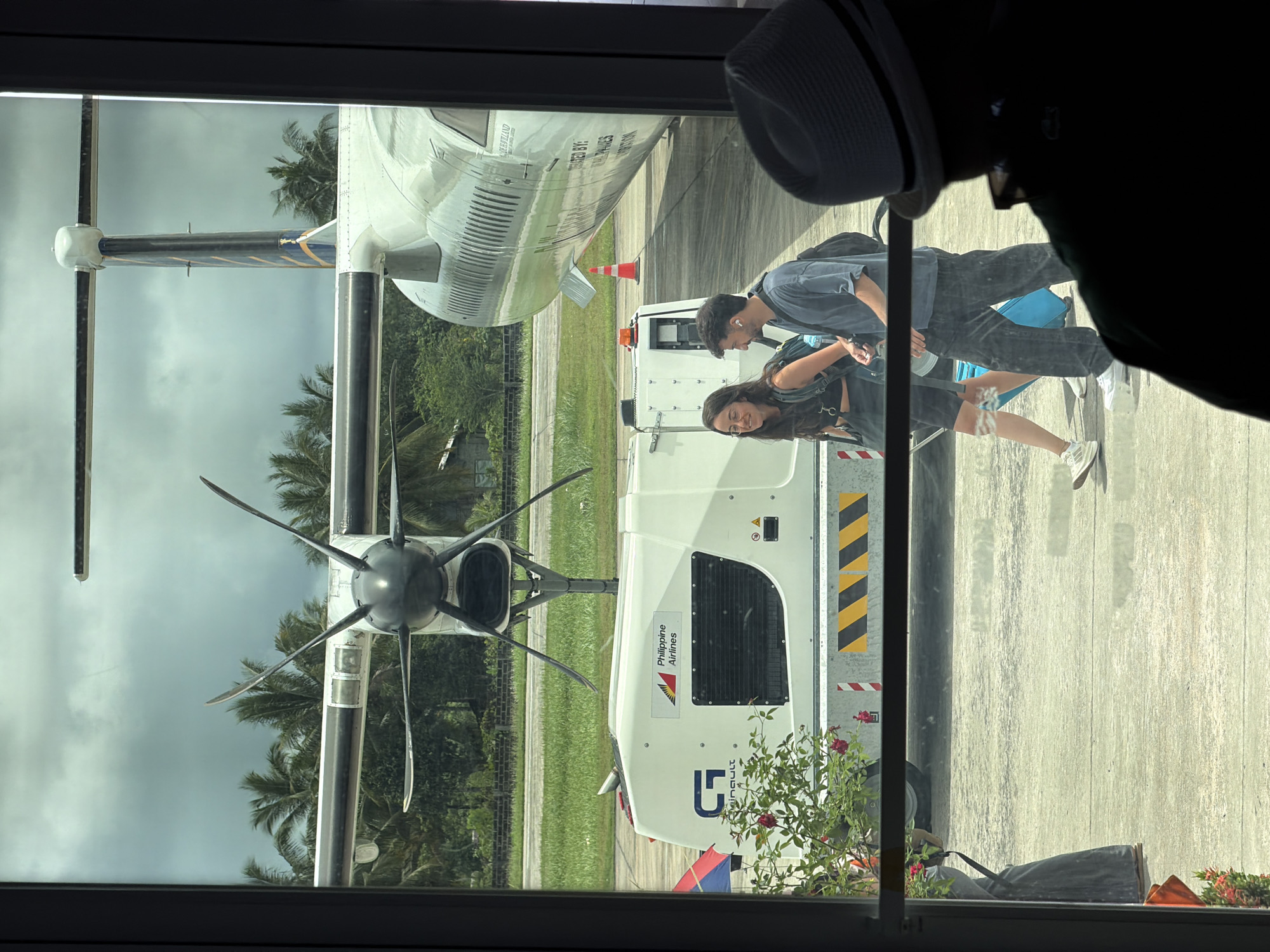 The image shows a view through a window in Del Carmen, Philippines, capturing an airplane on the tarmac and a couple walking towards it. The airplane has a visible propeller and is marked with "Philippine Airlines". The couple is casually dressed. A piece of airport equipment, possibly a generator, is positioned next to the plane, adorned with a "Philippine Airlines" logo and a yellow and black hazard stripe. Palm trees and overcast skies are visible in the background.