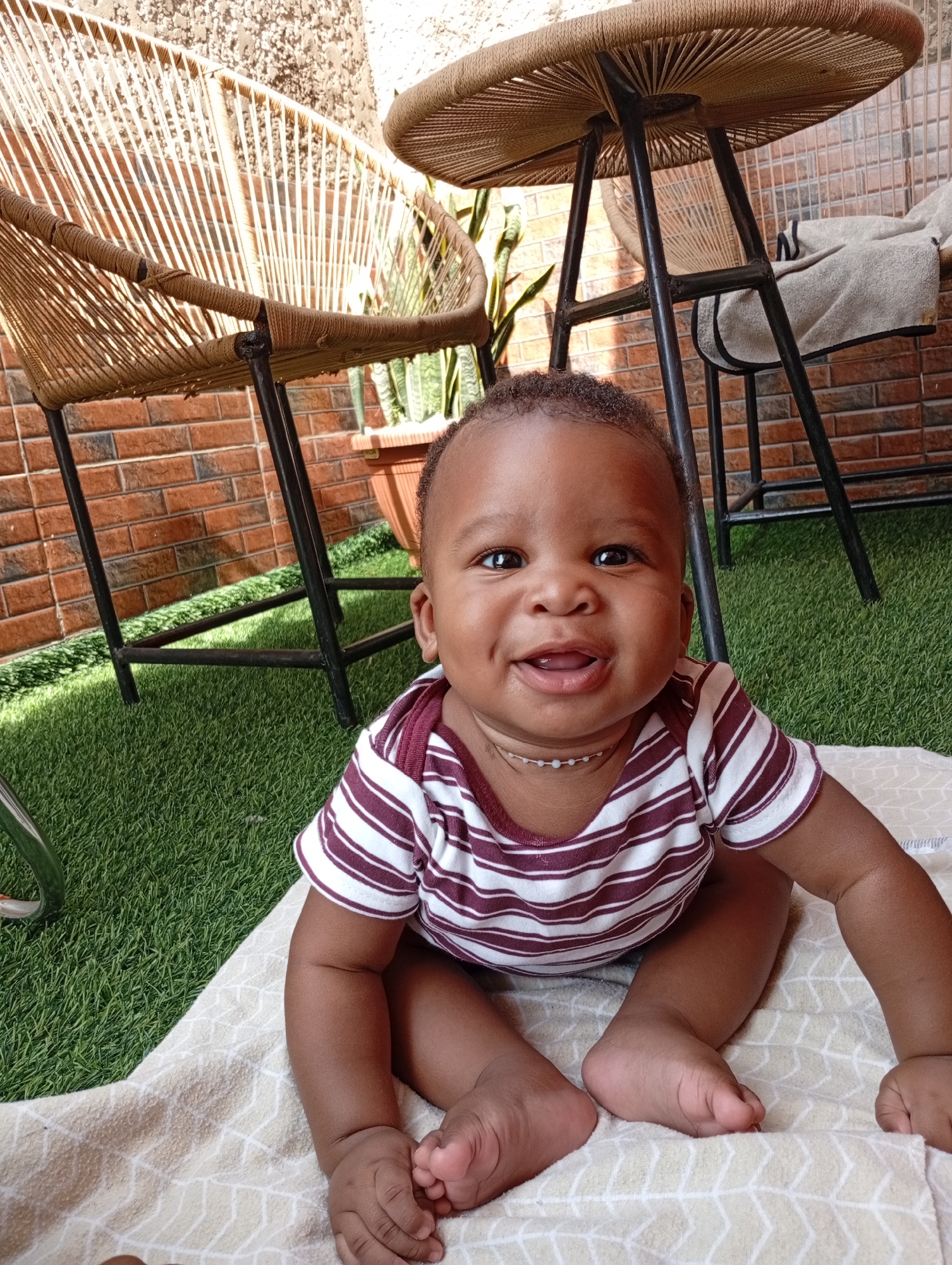 A dark-skinned baby with a round face, dark eyes, and a broad smile is sitting on a patterned blanket. The baby is wearing a white and maroon striped onesie and a delicate beaded necklace. The baby is on an outdoor patio with artificial grass underfoot. In the background, there are two woven chairs and a table with woven surfaces and black metal legs. A brick wall is visible behind the furniture. The overall setting suggests a residential outdoor space in Abuja, Nigeria.
