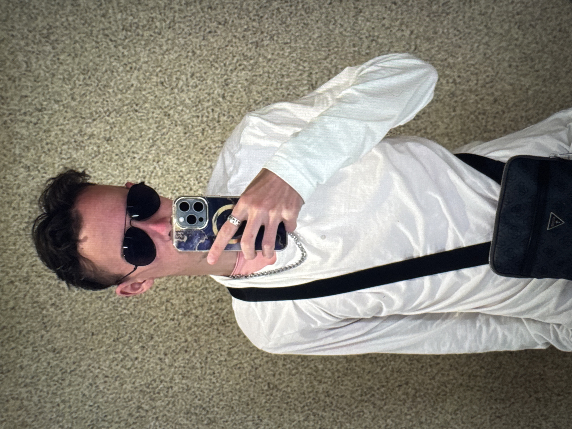 A young man, seen from the chest up, stands in front of a textured, neutral-colored wall. He is wearing a white long-sleeved shirt, aviator sunglasses, and a silver chain necklace. He is holding a smartphone with a marble-patterned case, taking a selfie. A black crossbody bag with a patterned strap is visible across his chest. His left hand, adorned with a ring, is holding the phone. The background appears to be an indoor wall with a speckled texture, possibly in a public space or residence in Porrentruy, Switzerland.