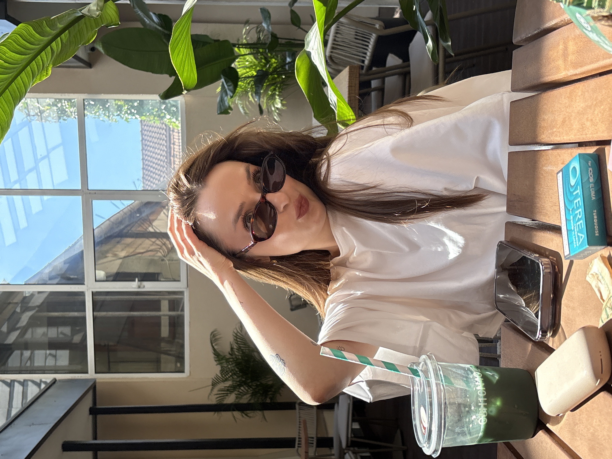 The image depicts a woman seated at a wooden table outdoors, in what appears to be a cafe or restaurant patio. She is wearing sunglasses and a loose-fitting white top, with her right hand resting on her head. On the table, there is a clear plastic cup filled with a green beverage and ice, a straw, a smartphone with a silver case, a beige electronic device, and a small packet of cigarettes. The background reveals an outdoor seating area with chairs and tables, surrounded by plants and a building with large windows. The overall lighting suggests it is daytime, possibly late morning or early afternoon. The location is identified as a cafe in Kragujevac, Serbia.