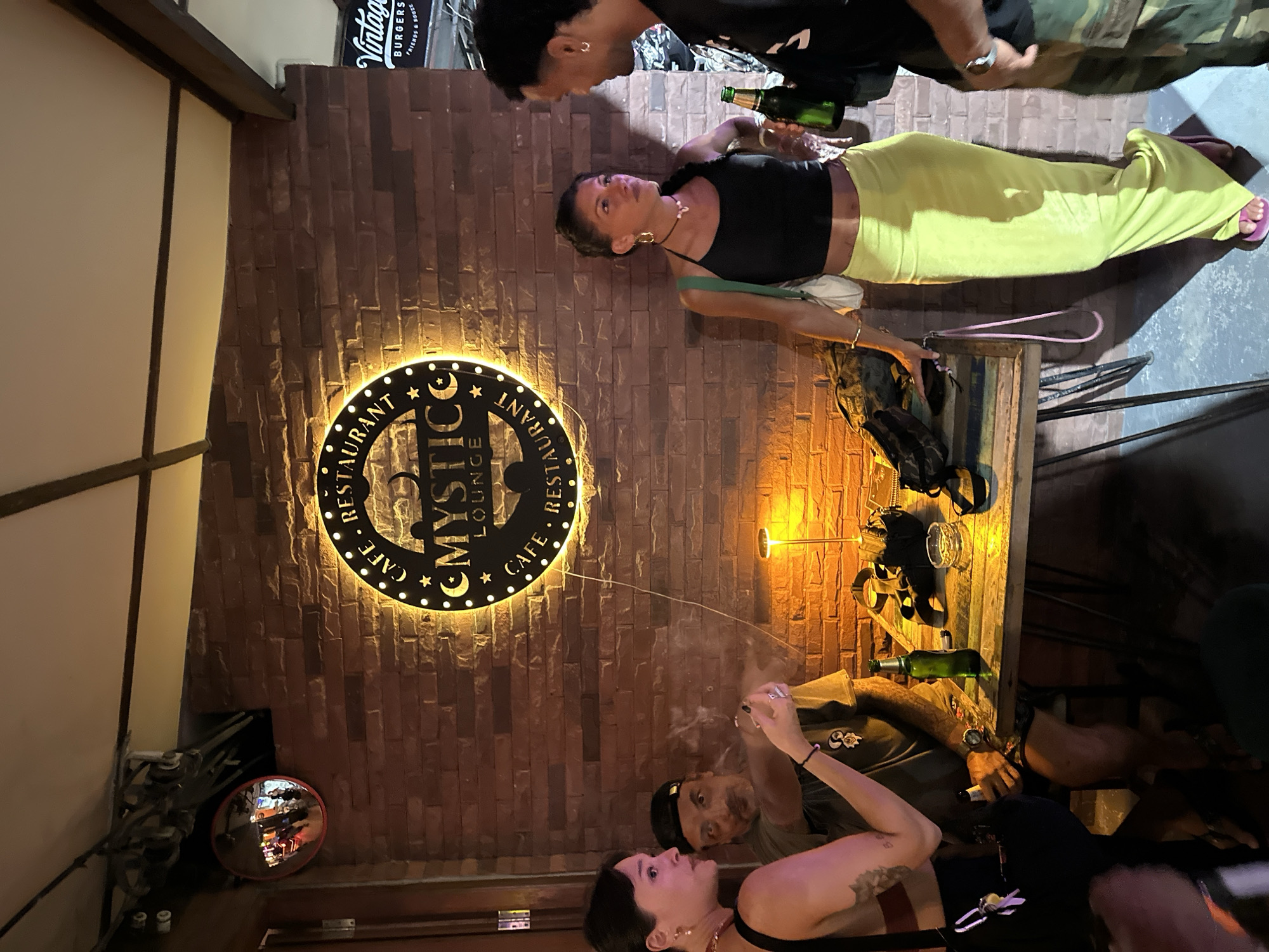 The image shows a group of people in what appears to be an indoor restaurant or lounge setting. A woman in a black top and bright yellow pants is standing and leaning against a brick wall, holding a drink. Another woman is seated in the foreground, looking up. A man is visible behind her, and another man is standing behind the woman in yellow. There is a round, illuminated sign on the brick wall with text that reads "MYSTIC CAFE RESTAURANT LOUNGE". Several bottles and glasses are present on a wooden surface. The lighting suggests it is nighttime or indoors with artificial lighting. The overall atmosphere seems casual and social. A sign with "Vintage Burgers" is also visible in the background. The location context is Koh Phangan, Thailand.