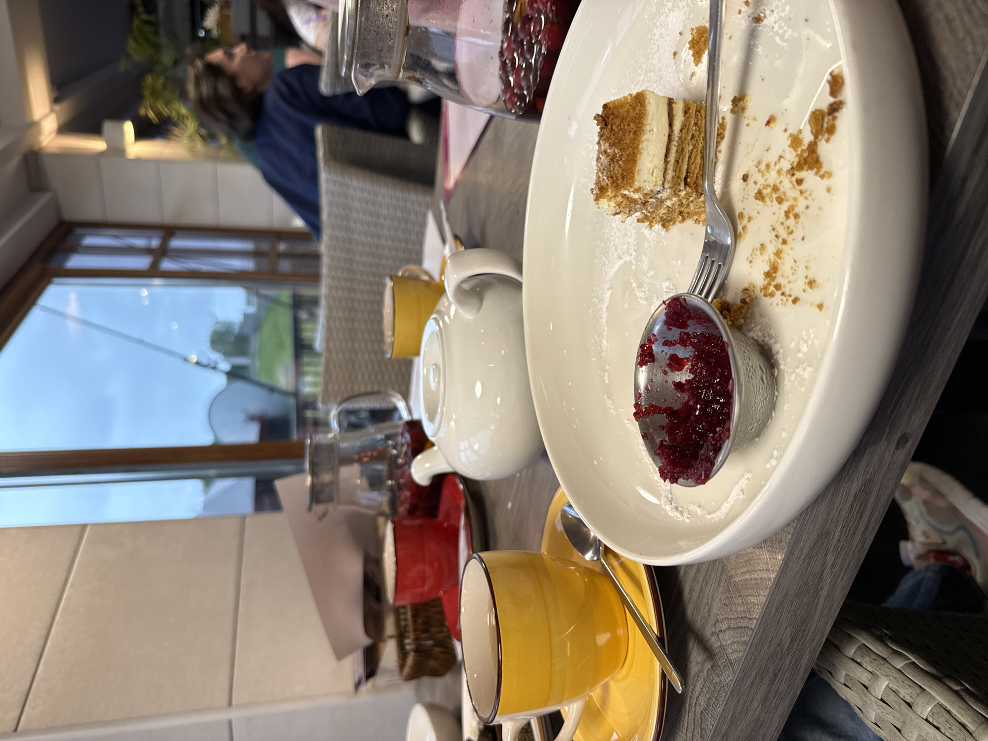 The image depicts a close-up view of a table setting in what appears to be a cafe or restaurant in Veliky Novgorod, Russia. In the foreground, a white plate holds a partially eaten slice of layered cake dusted with powdered sugar, a small metal ramekin filled with berry jam, and a fork. To the left, a yellow teacup sits on a matching saucer, with a spoon resting on the saucer. Behind this, a red teacup and saucer are visible. Further back, a white teapot and a glass carafe containing a red liquid with berries are present. In the background, through a large window, a glimpse of the exterior can be seen, with greenery and a cloudy sky. A person wearing a dark blue jacket is seated at another table, partially visible. The overall ambiance suggests a casual dining or tea experience.