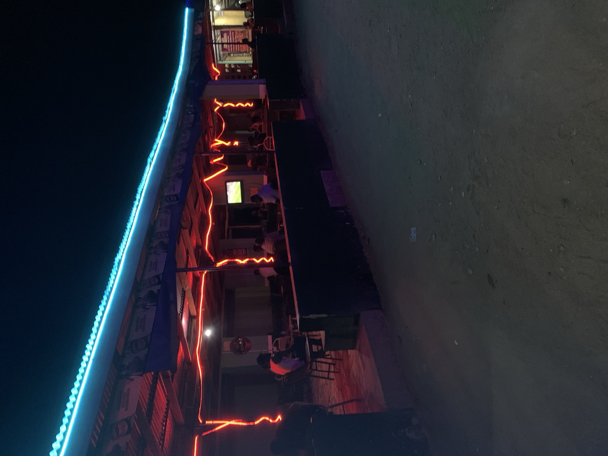 The image shows an outdoor bar at night in New Stadium, Nigeria. The bar's roof is lined with blue lights, and red lights zig-zag down the side of the building. People are seated at tables inside. A television is visible, and posters are affixed to the building. The ground outside the bar appears to be dirt or gravel.