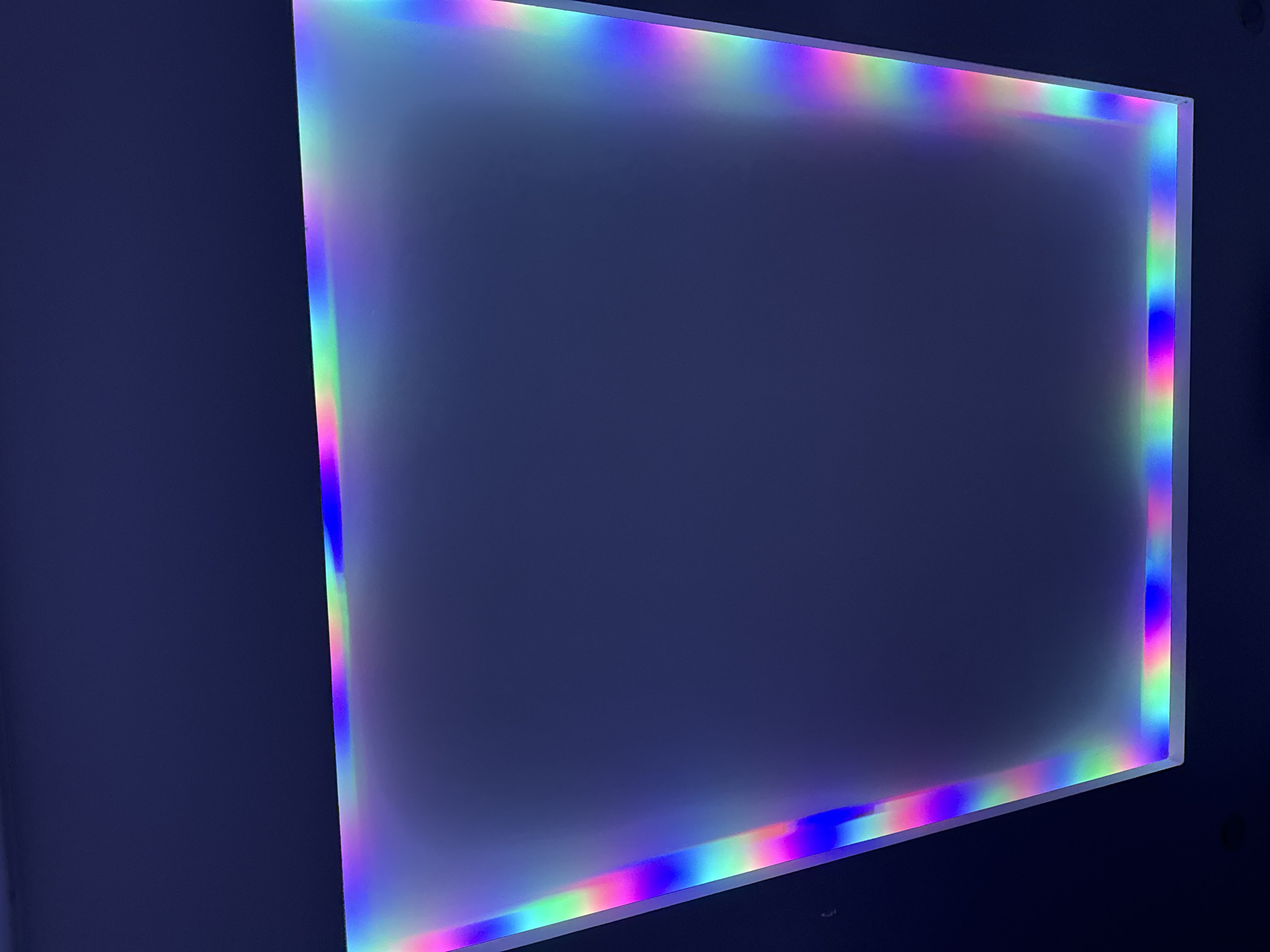 A rectangular light box with a rainbow colored edge is mounted on a dark blue wall. The light box dominates the scene, with its vibrant edge providing a sharp contrast to the somber background. The location is unspecified, and the only objects present are the light box and the wall. The absence of people, actions, or interactions suggests a static, inanimate scene. The environment consists solely of the indoor space with the dark blue wall. Mũi Né, Vietnam is not discernable from the image provided.