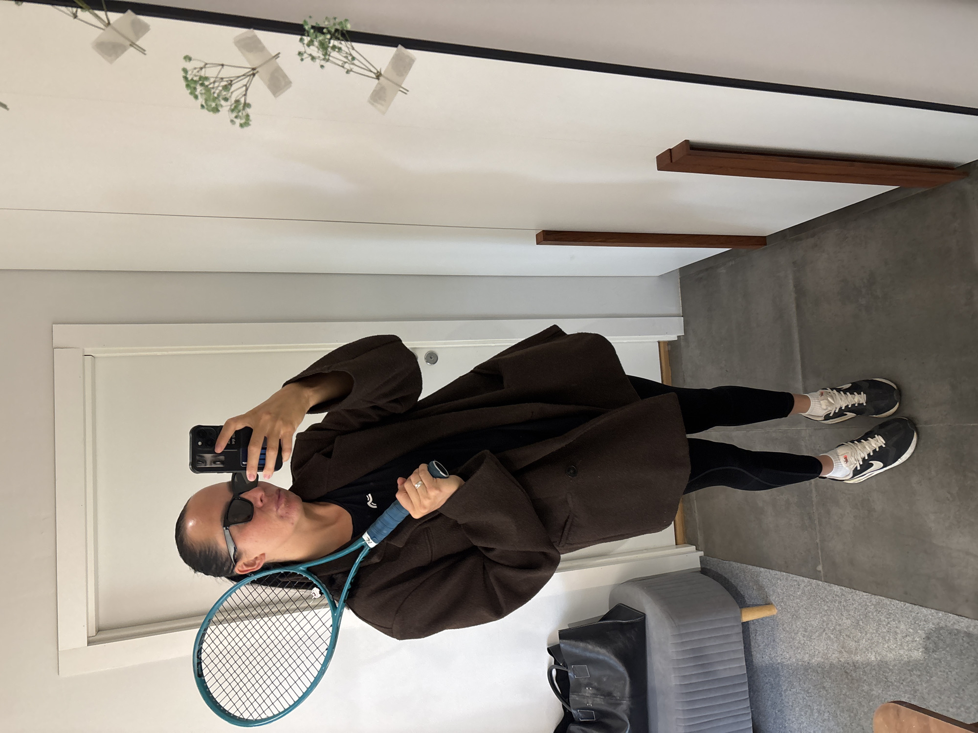 A person stands indoors, facing a mirror and holding a blue tennis racket in their left hand and a black smartphone in their right hand, apparently taking a selfie. They are wearing a brown overcoat, black leggings, a black t-shirt, and black and white sneakers. The person is also wearing sunglasses. In the background is a white wall with a doorframe, a gray floor, a gray tufted bench and a black leather tote bag. Tape and plant cuttings are visible on the mirror surface. Two wooden horizontal bars are visible on the wall. The location in the city of Chaikovsky, Russia, cannot be determined from the image.