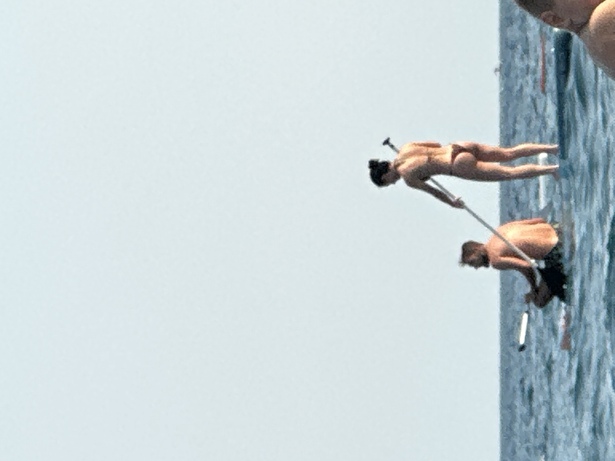 In the city of Boreti, Montenegro, two individuals are on paddleboards in the sea. The person on the right is a woman wearing a bikini, standing and holding a paddle. The person on the left is a man, sitting and also holding a paddle. The water is blue and choppy. The sky is clear and pale blue. A third person's head is visible in the foreground on the right.
