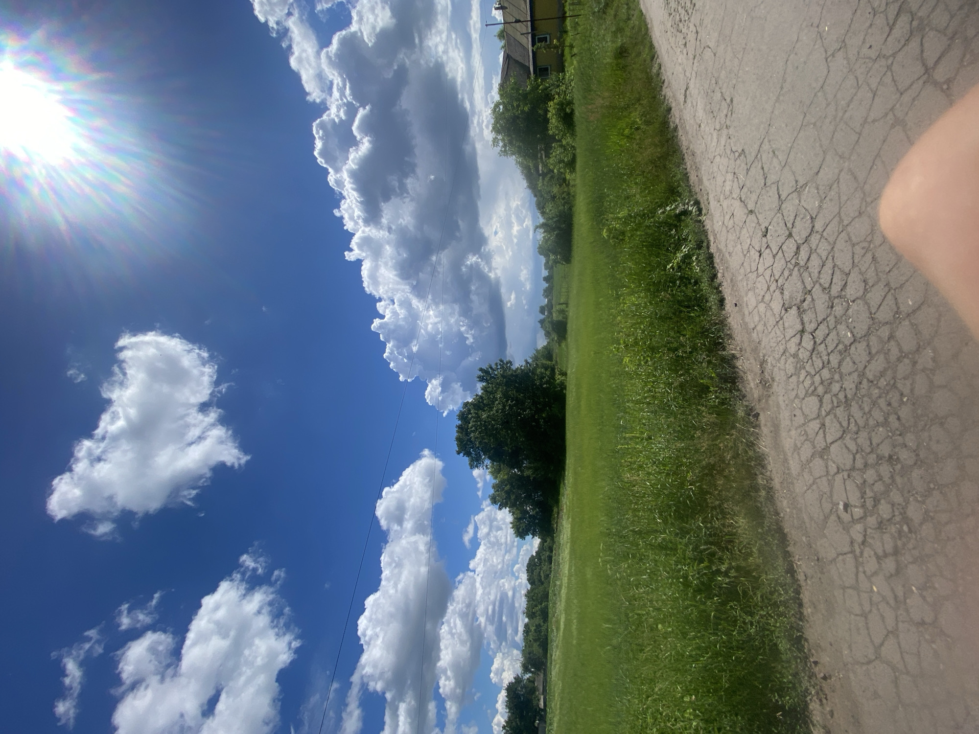 The image depicts a rural scene in Ustyvytsia, Ukraine, on a sunny day. In the foreground, a cracked asphalt road occupies the bottom right portion of the frame. Adjacent to the road is a lush green field of tall grass. The middle ground is characterized by more greenery, including a cluster of mature trees. To the right of the trees, a yellow house with a dark roof is visible. The background consists of a clear blue sky dotted with large, fluffy white clouds. A bright sun, partially obscured by lens flare, shines from the upper right corner. Overhead power lines are faintly visible stretching across the sky. The overall environment is that of an open, natural landscape with signs of human habitation.