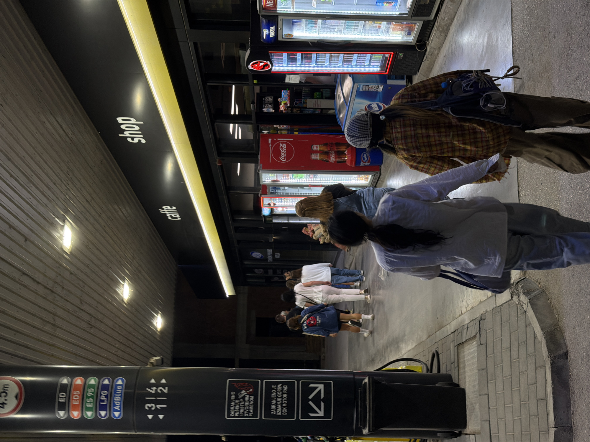 The image depicts a street scene at night, likely a gas station, with a shop and cafe area. The location is identified as Zvornik, Bosnia and Herzegovina.
In the foreground, two individuals are walking away from the camera. The person on the left wears a light-colored long-sleeved shirt and light-colored pants, carrying a dark bag over their shoulder. The person on the right is wearing a plaid shirt, dark pants, and a backpack, with a checkered hat on their head.
A group of four women is walking ahead of them, approaching the shop. They are dressed casually, with one in a blue jacket, another in a white top and light pants, and two more in white tops and jeans.
The environment features a gas pump on the left side of the image, with various fuel types indicated on a vertical panel. The background shows the facade of a building with large windows, displaying rows of illuminated beverage coolers. Signs for "caffe" and "shop" are visible above the entrance. The ground is paved with asphalt and some cobblestone. The lighting is artificial, primarily from the shop's interior, the coolers, and overhead lamps, creating a night-time ambiance.