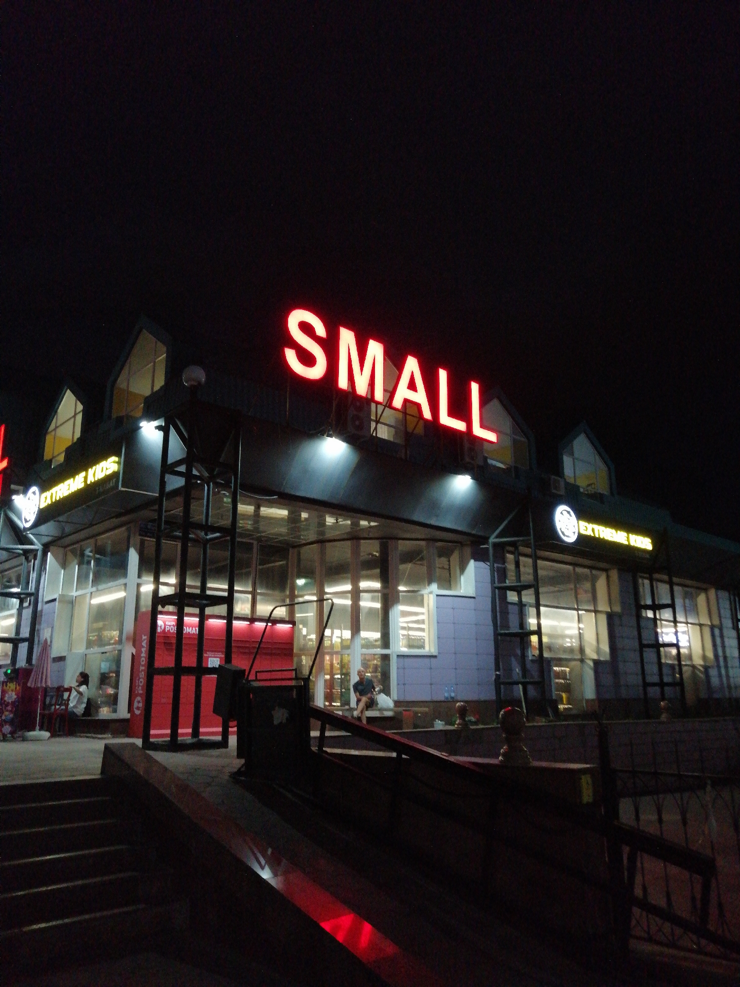 The image shows a neon sign that reads "SMALL" in red letters on the facade of a building at night. Below the "SMALL" sign, there are two smaller signs that read "EXTREME KIDS" in yellow letters, one on each side of the building. The building appears to be a commercial establishment with large windows and a modern architectural design. In front of the building, there is a red post office box labeled "POSTOMAT." There are a few people visible in the image, some sitting on benches and others walking. The scene is set in the city of Pavlodar, Kazakhstan. The lighting suggests it is nighttime, with artificial lights from the building illuminating the area.