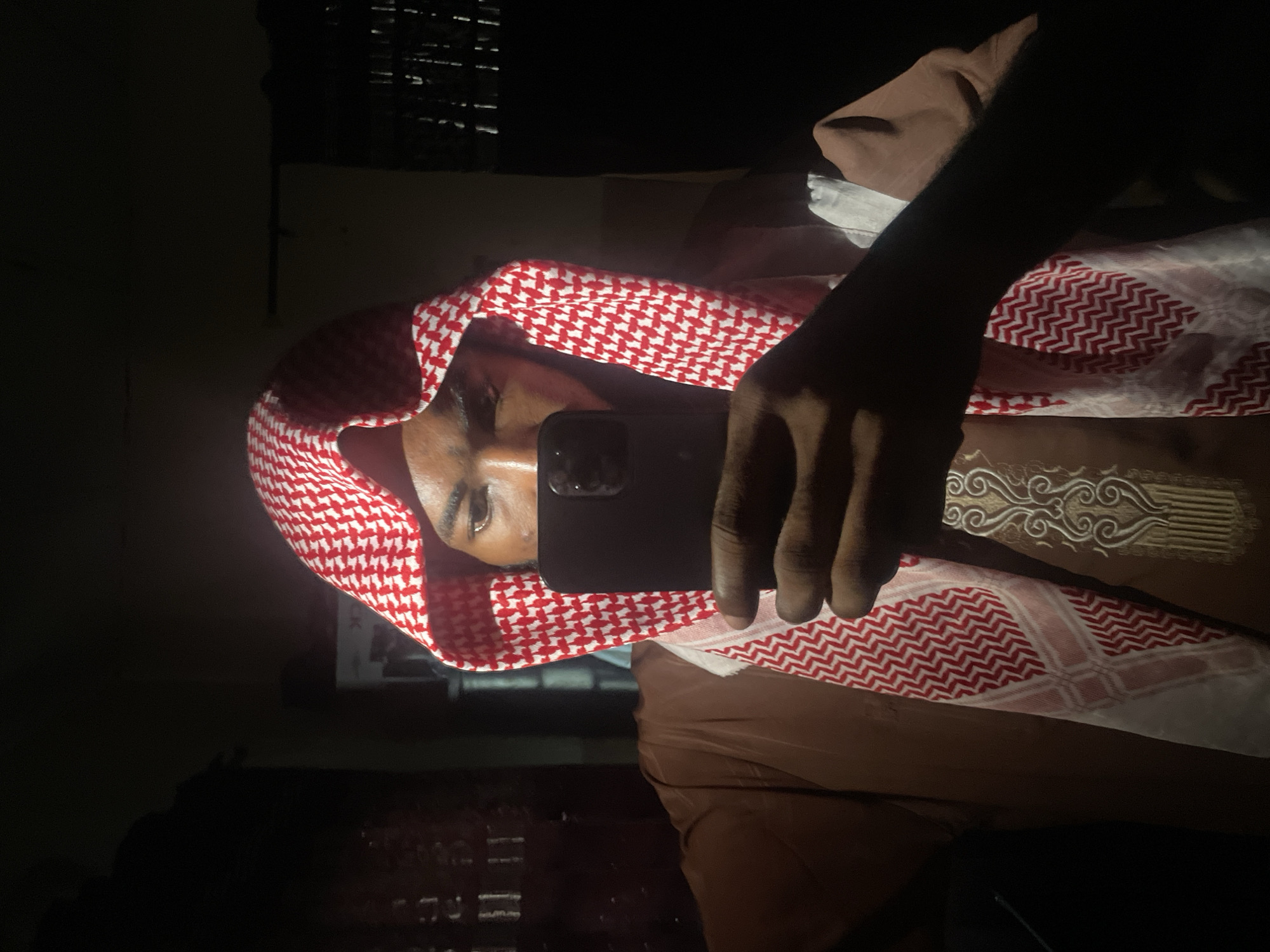A man wearing a red and white checkered keffiyeh is visible, holding a smartphone in his right hand. The phone obscures part of his face. He is wearing a brown garment, possibly a traditional robe, with ornamental patterns on the sleeve. The environment is dimly lit. A window with horizontal blinds can be discerned in the background. The location, Lokoja, Nigeria, is based on provided instructions.