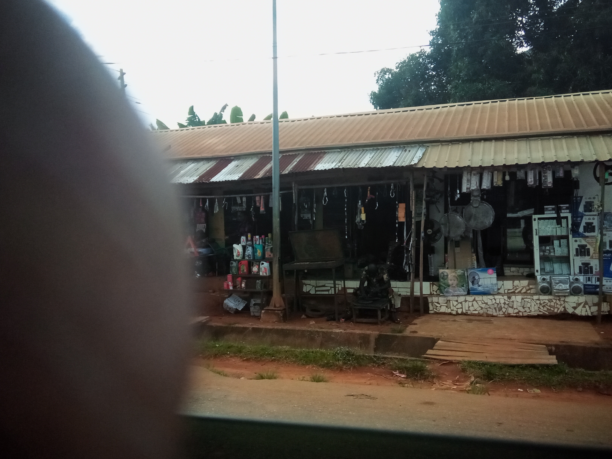 The image depicts a roadside market stall in Nibo, Nigeria. The stall has a corrugated metal roof supported by wooden poles and is lined with various goods. On the left side, shelves display an assortment of colorful plastic bottles, likely containing motor oil or lubricants. In the center, a dark, flat-screen television or monitor is visible, resting on a table. To the right, more shelves are stacked with electronic appliances, including what appear to be speakers and possibly a radio or DVD player. A person, dressed in dark clothing, is seated on a stool to the right of the central table, partially obscured by shadows. The ground in front of the stall is a mixture of dirt, grass, and a paved pathway. Wooden planks are laid across a section of the pathway. The background shows trees and a bright sky. The overall scene suggests a typical informal commercial setting.