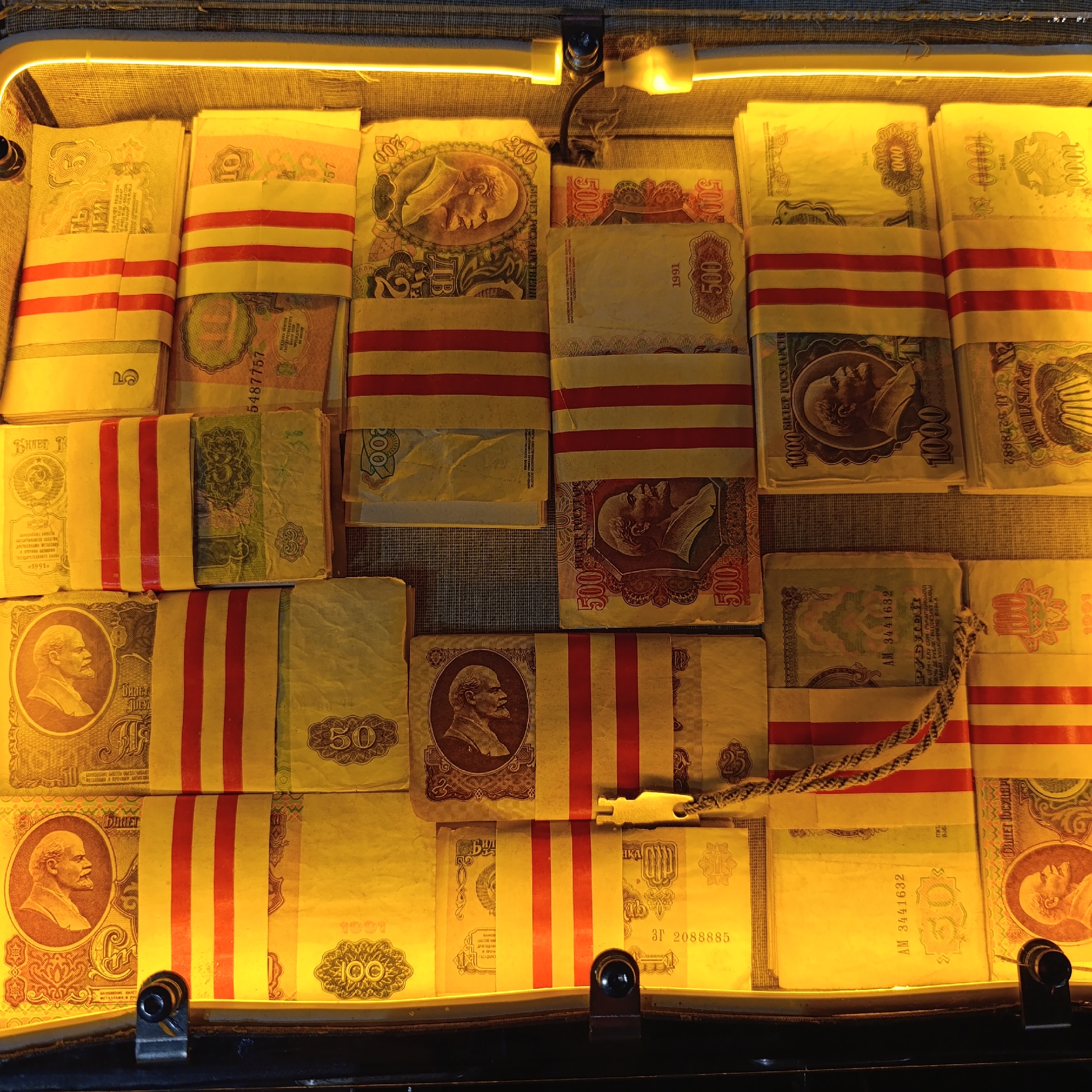 This image captures a curated display of numerous Soviet-era banknotes, arranged neatly in bundles within what appears to be a lighted exhibition case or suitcase. The currency is predominantly secured with distinctive red and white striped bands.
The subjects are stacks of various denominations, including 1, 3, 5, 10, 50, 100, 200, 500, and 1000 rubles. Many of the banknotes feature the portrait of Vladimir Lenin, a common motif on Soviet currency. A 200-ruble note in the upper right displays a different, possibly allegorical, female figure.
The activity depicted is the presentation of these historical financial instruments, likely as part of a museum collection or numismatic exhibit. The uniform arrangement and securing of the bundles suggest a careful and organized display.
Notable details include the warm, yellowish glow emanating from a strip light positioned along the top edge of the case, highlighting the currency. Visible dates on the banknotes include 1991 and 1992, indicating currency from the late Soviet period and its immediate aftermath. Visible text includes "БИЛЕТ ГОСУДАРСТВЕННОГО БАНКА" (State Bank Note), "РУБЛЕЙ" (Rubles), and a warning about counterfeiting: "ПОДДЕЛКА БИЛЕТОВ ГОСУДАРСТВЕННОГО БАНКА ПРЕСЛЕДУЕТСЯ ПО ЗАКОНУ" (Forgery of State Bank Notes is prosecuted by law). Several serial numbers like "5487757", "AM 3441632", and "31 2088885" are also legible. The setting is indoors, typical for an exhibition, and the historical context aligns with Verkhnyaya Pyshma, Russia, as a location for such a display of national heritage.