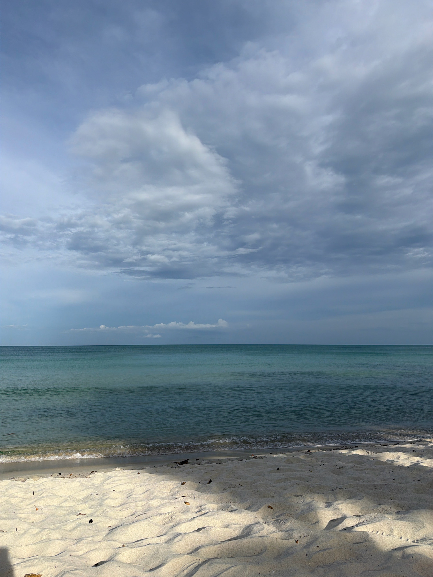 A wide-angle, eye-level shot captures a serene beach scene on Ko Samui, Thailand. The foreground is dominated by pristine white sand, subtly textured with ripples and casting soft shadows. A scattering of small leaves and debris dots the sand. The gentle edge of the turquoise ocean laps at the shore with a thin line of white foam, creating a subtle wave pattern. The middle ground consists of the calm, clear water, transitioning from a lighter turquoise near the shore to a deeper blue as it recedes towards the horizon. The horizon line is sharp and straight, dividing the water from the sky. The sky above is dramatic, filled with a vast expanse of cumulonimbus clouds in varying shades of grey and white. Patches of pale blue sky are visible between the clouds, hinting at the possibility of sunshine breaking through. The overall atmosphere is tranquil and natural, suggesting a peaceful moment on a tropical coastline.