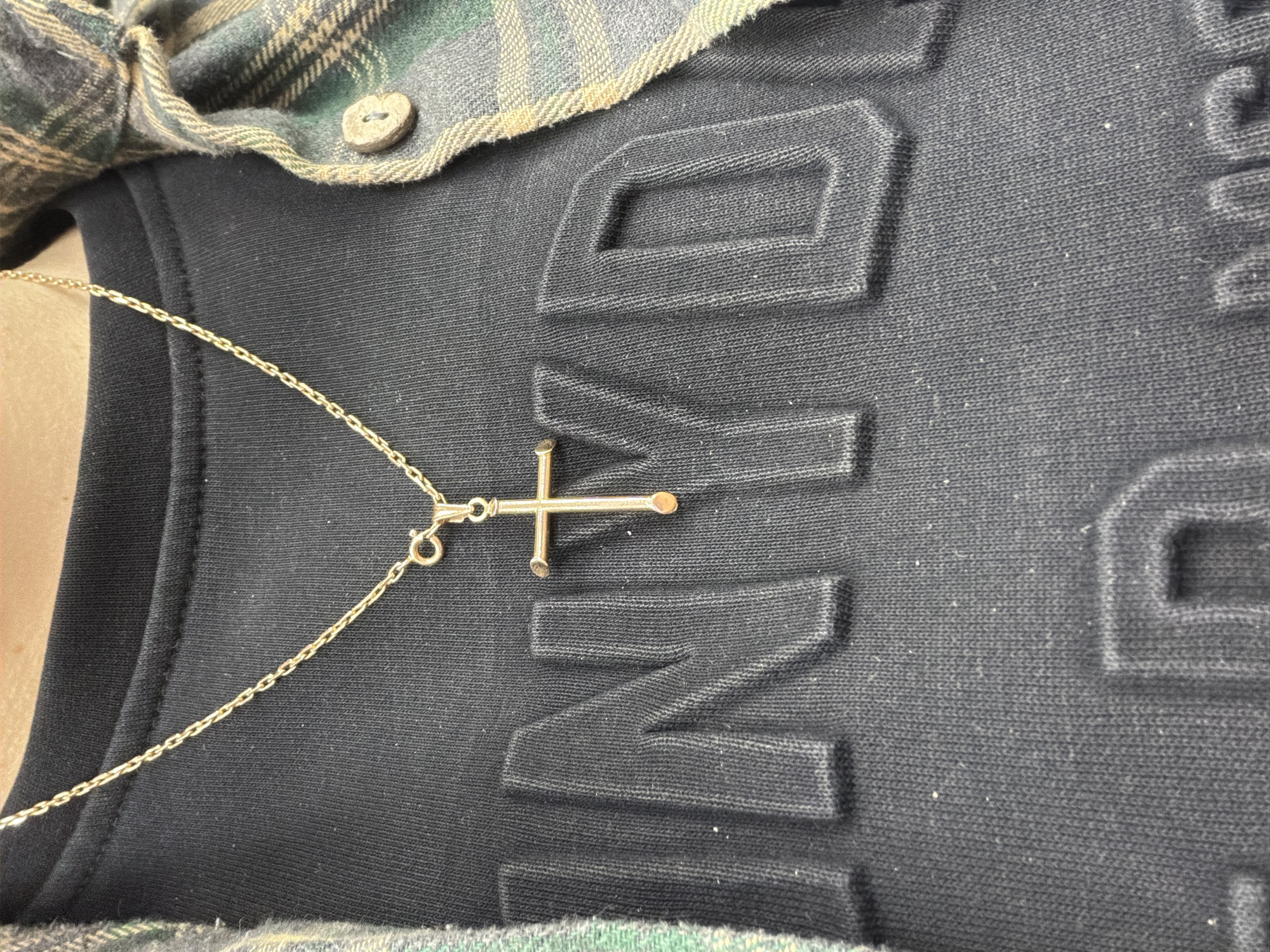 A close-up shot captures the upper torso of an individual in Sochi, Russia, wearing casual attire. A dark, possibly black, t-shirt with embossed text is visible, displaying parts of letters that appear to spell "VID" and "N", hinting at a word such as "DAVID" or "DIVISION". Over the t-shirt, a plaid flannel shirt in shades of green, black, and beige is worn, with a natural-colored, possibly wooden, button visible near the collar. A delicate gold-colored chain necklace with a small, simple cross pendant rests on the t-shirt. The cross has a minimalist design with rounded ends on its bars. The image focuses on the textures of the fabrics and the detail of the jewelry, with no specific activity taking place. The lighting is even, revealing the fine weave of the t-shirt and the slight fuzz on its surface.