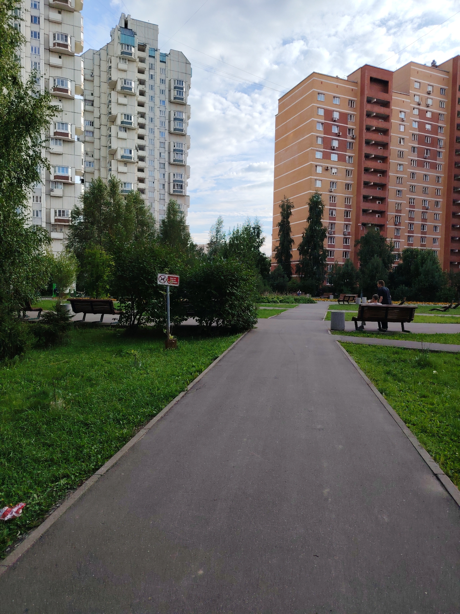 The image depicts a residential area in Zelenograd, Russia, featuring a paved walkway leading through a park-like setting with manicured grass and trees. Two multi-story apartment buildings dominate the background. The building on the left is a light-colored structure with numerous balconies and windows, while the building on the right is an orange and brown brick building, also with many windows and balconies. Several park benches are visible, with two individuals, an adult and a child, seated on one of them. Other people are discernible in the distance. A sign with a red circular symbol and text, likely indicating a restriction, is posted near a bush. In the foreground, a strip of red and white tape is present on the grass near the edge of the walkway. The sky is partly cloudy.