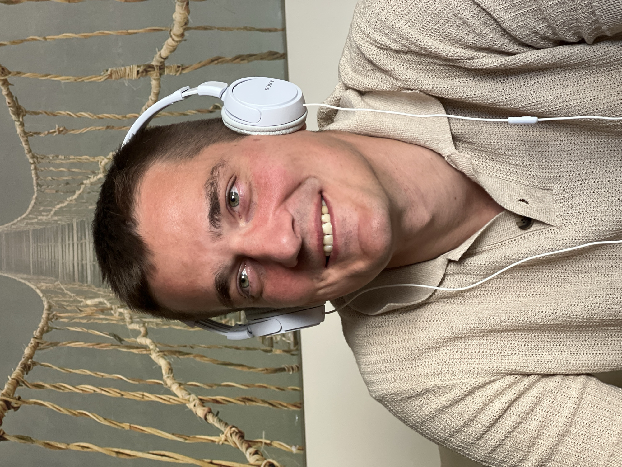 A man is wearing white Sony headphones. He is smiling. He is wearing a beige knitted shirt. A rope bridge is in the background, potentially in Budva, Montenegro.