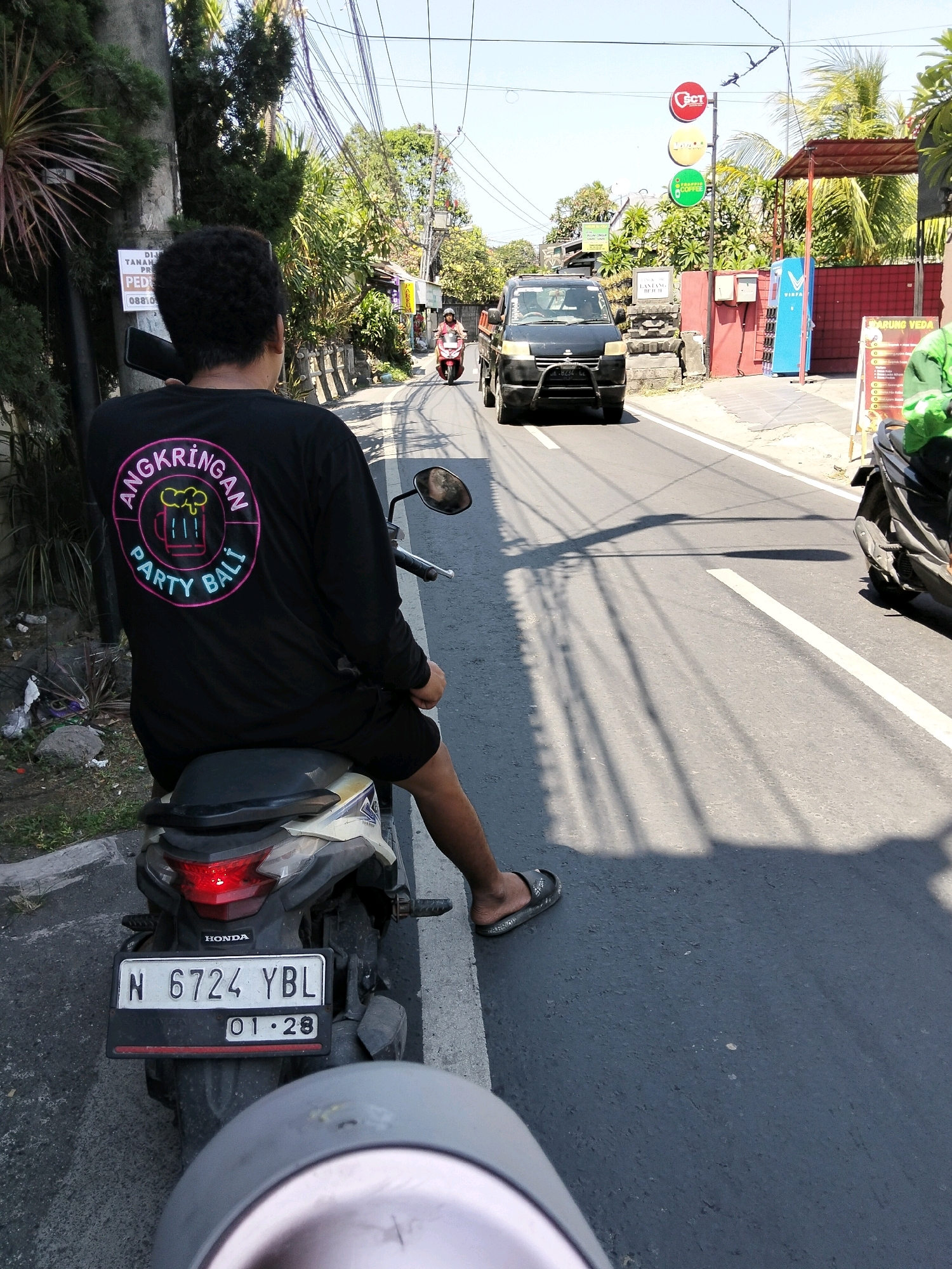 The image captures a street scene in Denpasar, Indonesia, likely on a sunny day with clear skies, indicated by the strong shadows. In the foreground, a person is seated on a motorcycle, facing away from the camera. They are wearing a black long-sleeved shirt with a circular graphic on the back that says "ANGKRINGAN PARTY BALI" with an illustration of a beer mug. The motorcycle's license plate reads "N 6724 YBL" and a sticker below it shows "01-28".
The motorcycle is parked on the side of a paved road, with a gray concrete curb next to it. Beyond the curb, there is a patch of dry vegetation and some debris. The road itself is marked with white lines and has tire tracks visible. In the midground, a black pickup truck is driving down the road. Further down the road, a red motorcycle with a rider is also visible.
On the right side of the road, there are buildings with various signs. One building has a red facade with a covered area and a blue dispenser. Signs on poles indicate businesses, one with circular logos colored red, yellow, and green, featuring text that appears to be "CCT," "JAVA," and "COFFEE." Another sign on a stone structure reads "Jalan Lang..." suggesting a street name. Overhead, a complex network of electrical wires crisscrosses the sky, a common sight in urban Indonesian environments. The overall impression is of a typical bustling street in an Indonesian city.