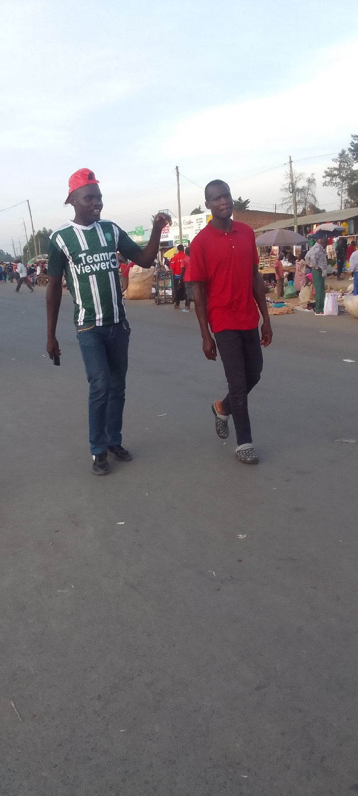 The image depicts two men walking on a paved road in what appears to be a busy market area in Kakunga, Kenya, during the daytime.
The man on the left is smiling cheerfully and looks towards his companion, with his right arm raised in a gesture. He is wearing a green and white striped short-sleeve shirt with "Team Viewer" written in white text across the chest. He also wears a red baseball cap backward, blue jeans, and dark shoes. He holds a dark object, possibly a phone, in his left hand.
The man on the right walks beside him, looking slightly to his left with a neutral expression. He is dressed in a red polo shirt, dark trousers, and dark clogs worn over light-colored socks.
The background reveals a bustling scene with numerous other people, market stalls, and structures lining the road, suggesting a vibrant commercial or public space. Buildings, utility poles, and distant trees are also visible. The sky is bright but softly lit, indicating it is daytime, possibly early evening or an overcast day.