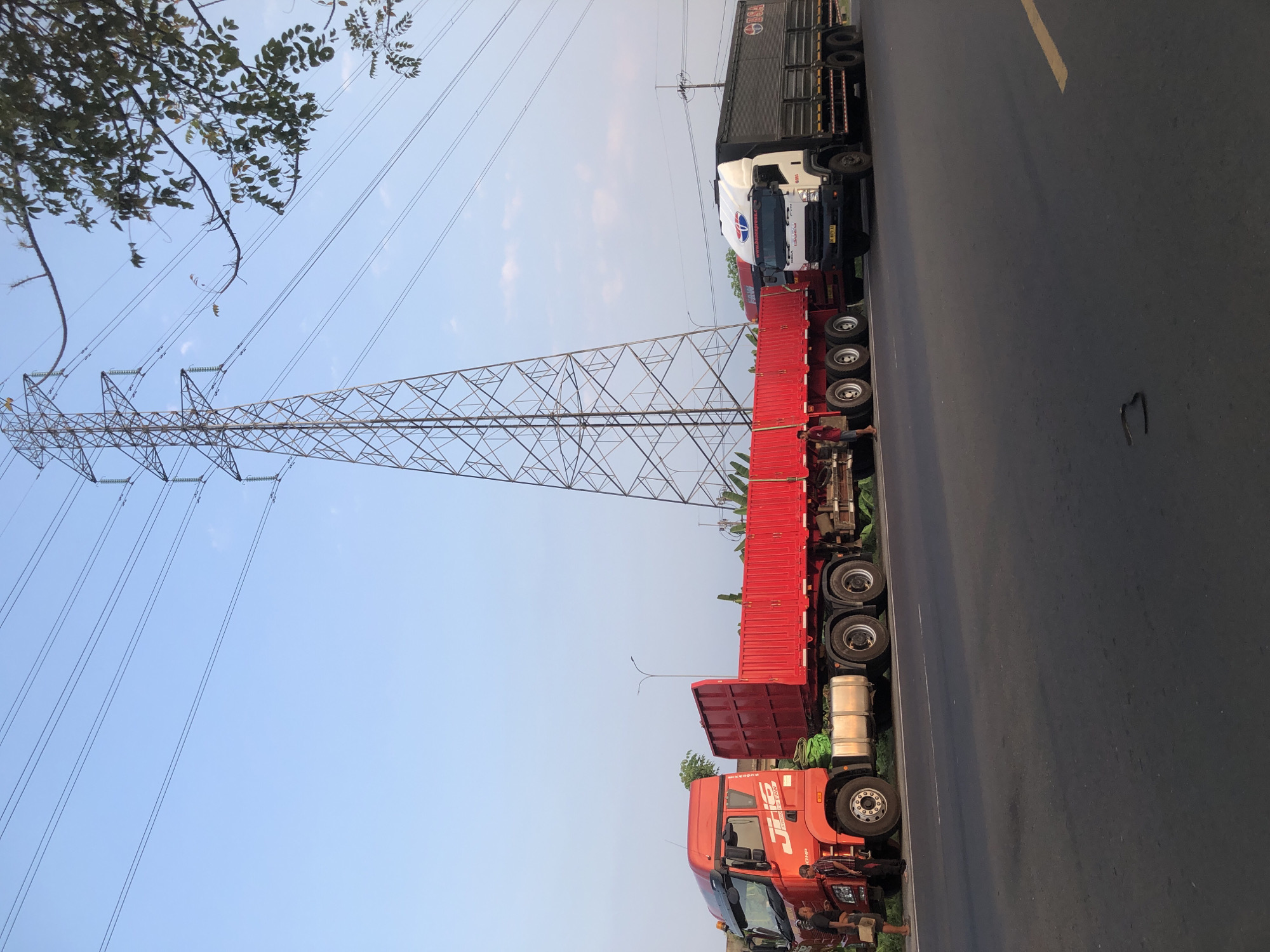 Two trucks are parked on the side of a road, next to a power line tower. The truck on the left is red and has a flatbed trailer. The truck on the right is white and black and has a covered trailer. Two men are standing near the red truck. The sky is clear and blue. The image appears to be taken in Pamotan, Indonesia.