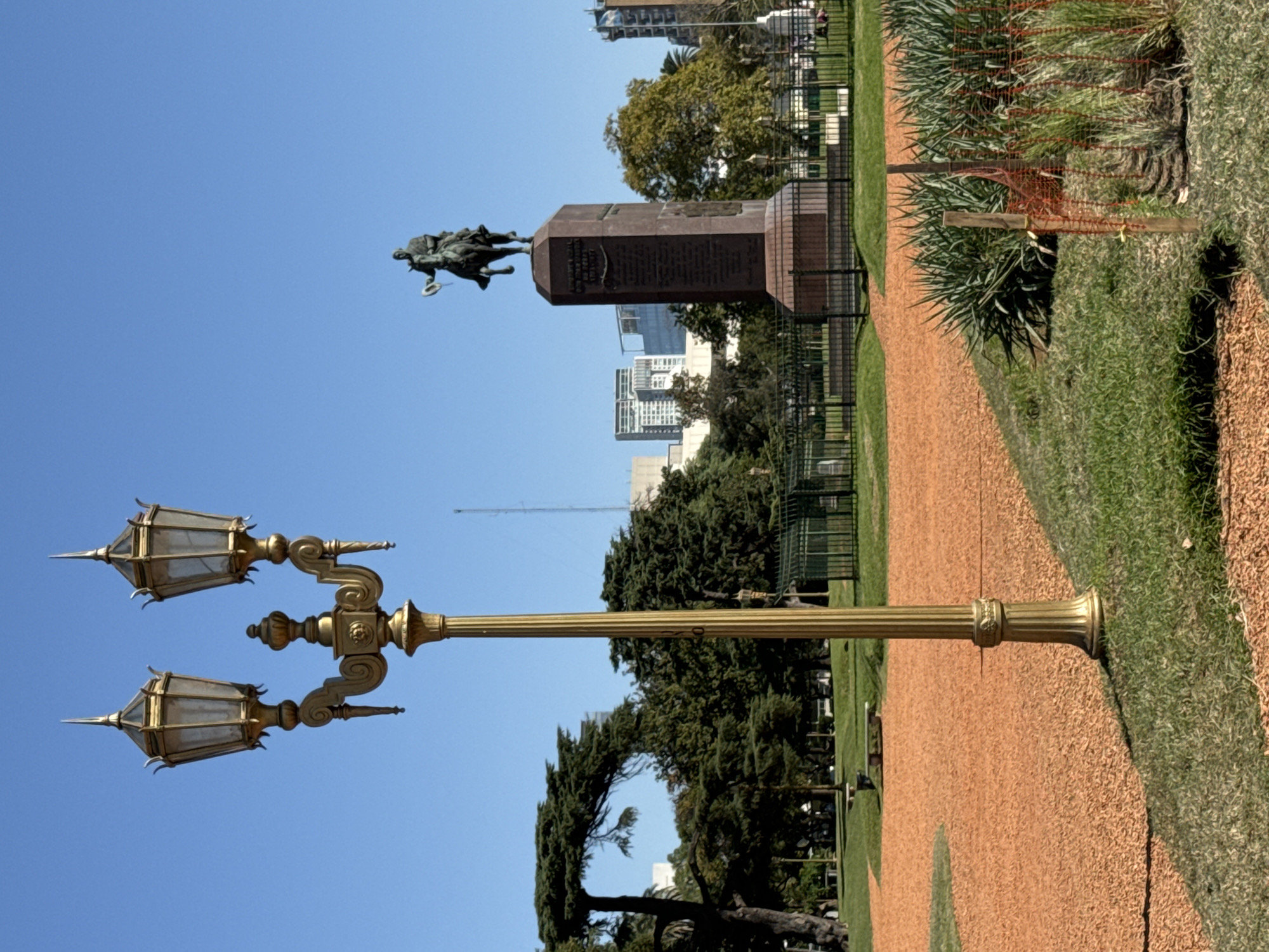 The image, taken in Buenos Aires, Argentina, presents a bright, sunny daytime view of a public park or garden. In the foreground, a reddish-brown gravel path curves through vibrant green grass. To the right of the path, a section of grass and foliage is marked by a wooden post with yellow ties and orange netting, suggesting a protected or newly planted area.
A tall, ornate golden lamppost with two elaborate lanterns dominates the left side of the frame. Its intricate design and classical styling stand out against the clear blue sky.
Further in the mid-ground, a prominent monument features a bronze equestrian statue atop a large, dark stone pedestal. The statue depicts a figure on horseback, possibly in a dynamic pose. Multiple lines of engraved text are visible on the monument's base, though the specific wording cannot be discerned due to distance. A black metal fence encloses the area around the monument.
The background reveals a cityscape with modern buildings interspersed among numerous green trees, indicating an urban setting. The sky is clear and blue, suggesting fine weather and bright sunlight. The scene captures a blend of historical memorial and contemporary parkland within a bustling city environment.
