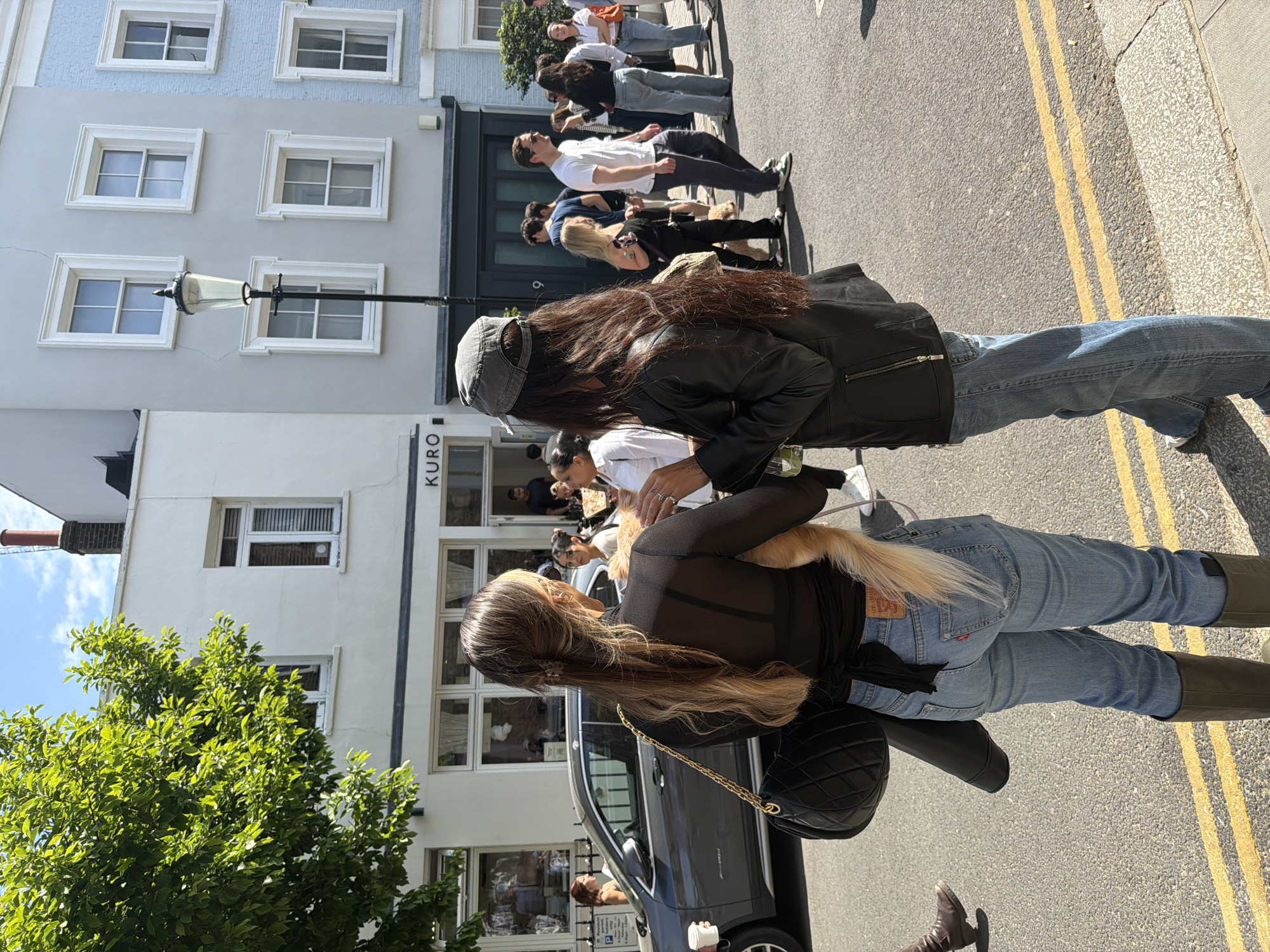 The image depicts an outdoor scene in London, United Kingdom, on a sunny day. Two women are in the foreground. One is wearing a black leather jacket, a grey cap, and jeans. The other wears a black top, light blue jeans, and high boots; she is holding a small dog. Behind them, a line of people is waiting outside a building labeled "KURO". Cars are parked alongside the road with double yellow lines, indicating parking restrictions. Buildings of varying colors and architectural styles are visible in the background, suggesting an urban setting in a commercial area. Trees and foliage are also visible, adding to the urban landscape. The overall atmosphere suggests a bustling street scene in London.
