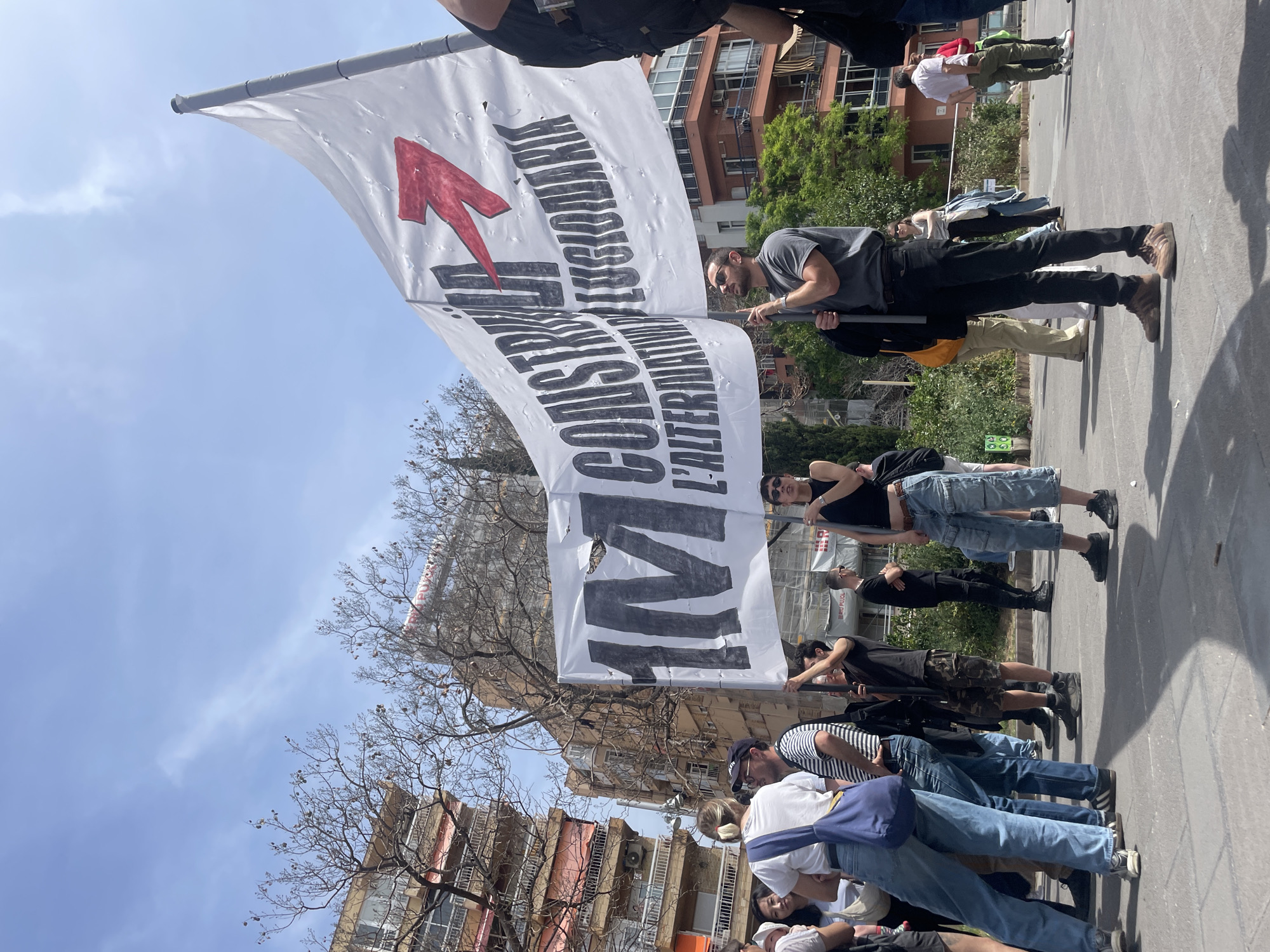 A large white flag, held upright by several people, dominates the center of the frame. The flag is emblazoned with a prominent red arrow pointing upwards and to the left, alongside large black text that reads: "M CONSTRUIM L'ALTERNATIVA". This phrase translates from Catalan to "We build the alternative." The flag shows signs of wear, with several small holes scattered across its surface.
A group of people, mostly adults, are gathered on a street, with some actively holding the flagpole. They are dressed in casual attire, including t-shirts, jeans, shorts, and caps. One person holding the flag wears sunglasses and a grey t-shirt.
In the background, typical urban residential buildings with multiple stories, balconies, and windows line the street. Some greenery from trees and bushes is visible, with some trees appearing bare-branched. Scaffolding is present on one of the buildings. The ground is paved, suggesting a street or public square.
The scene unfolds under a bright, clear blue sky with a few wispy clouds, indicating sunny weather during the day, likely mid-morning to afternoon, as suggested by the strong shadows. The overall setting is an outdoor, urban environment, capturing what appears to be a public demonstration or gathering in Barcelona, Spain.