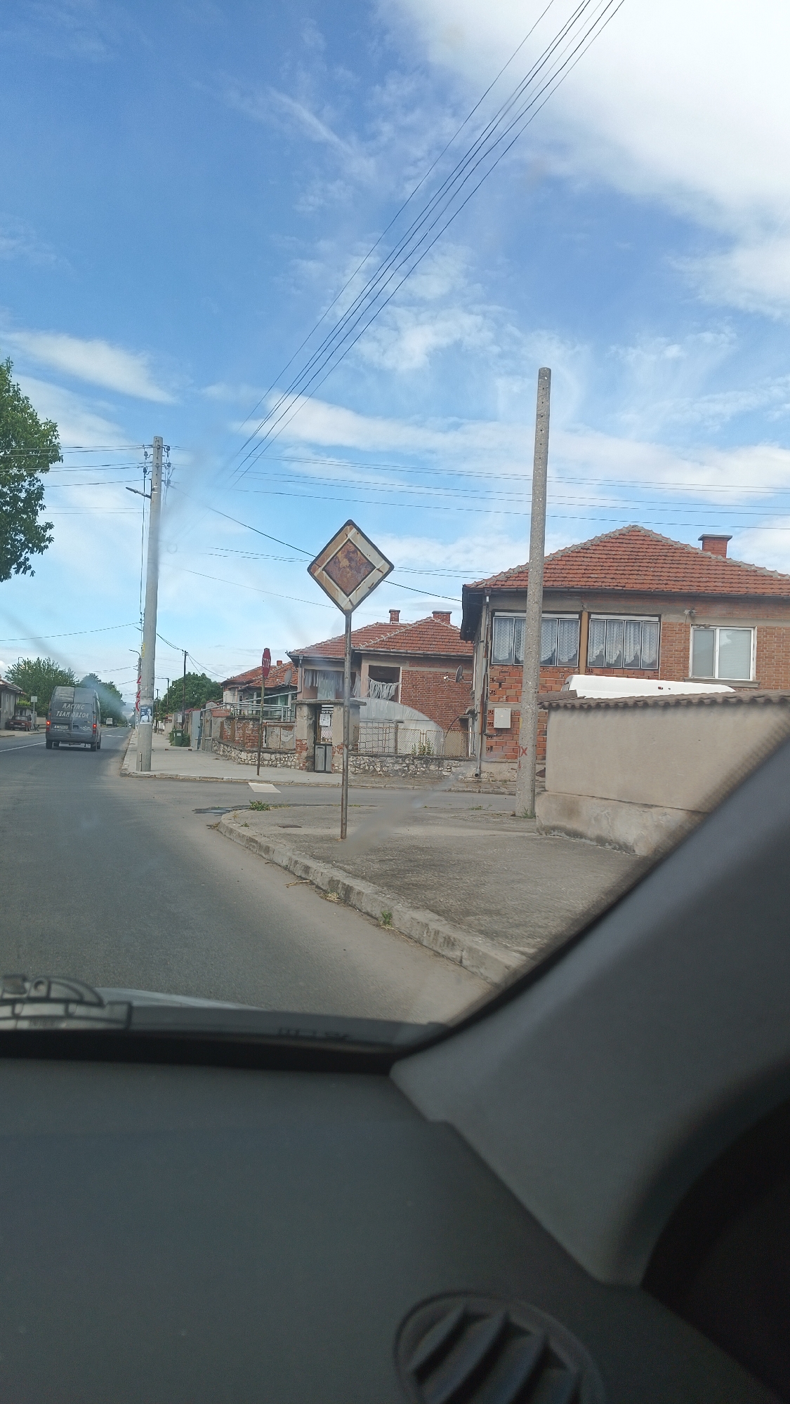The scene, taken from inside a vehicle, shows a street intersection in Milevo, Bulgaria. A weathered diamond-shaped traffic sign stands at the corner, indicating a yield or intersection ahead. Behind the sign are residential buildings with red tile roofs and brick facades. An adjacent concrete wall is partially visible. A vehicle is visible in the distance. Overhead power lines are strung across the sky, which is blue with scattered clouds. Part of the dashboard and a vent are visible in the foreground.