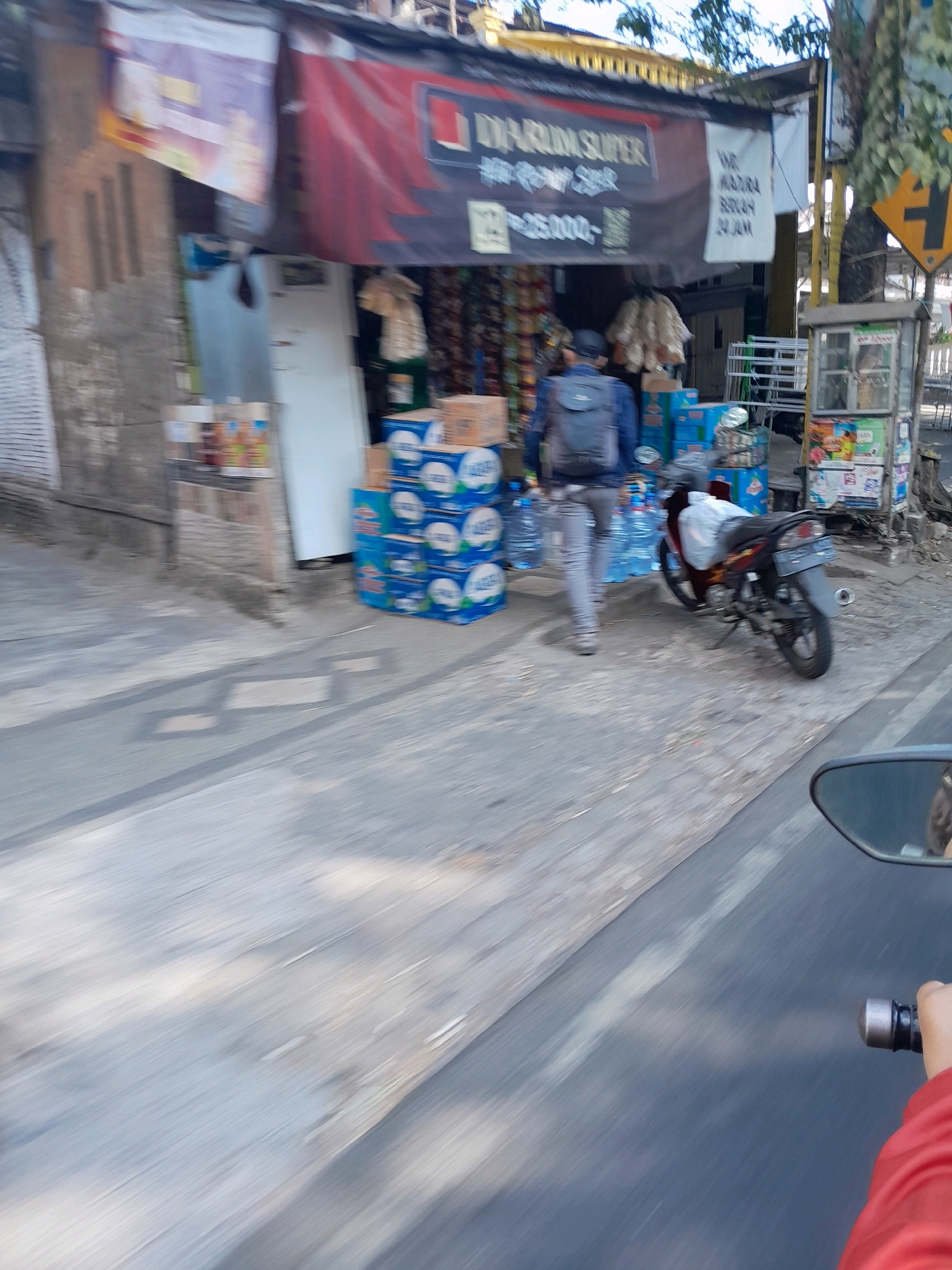 The image depicts a street scene in Ngadilangkung, Indonesia, with a small roadside shop as the central focus. A person wearing a backpack is walking away from the shop, which displays banners with text in Indonesian, including "DINARLIN SUPER" and what appears to be a price of "Rp25.000,-". Stacks of bottled water are visible in front of the shop, and a motorcycle is parked nearby. The environment is urban, with paved roads and building facades. The overall atmosphere suggests a typical day in a local Indonesian town.