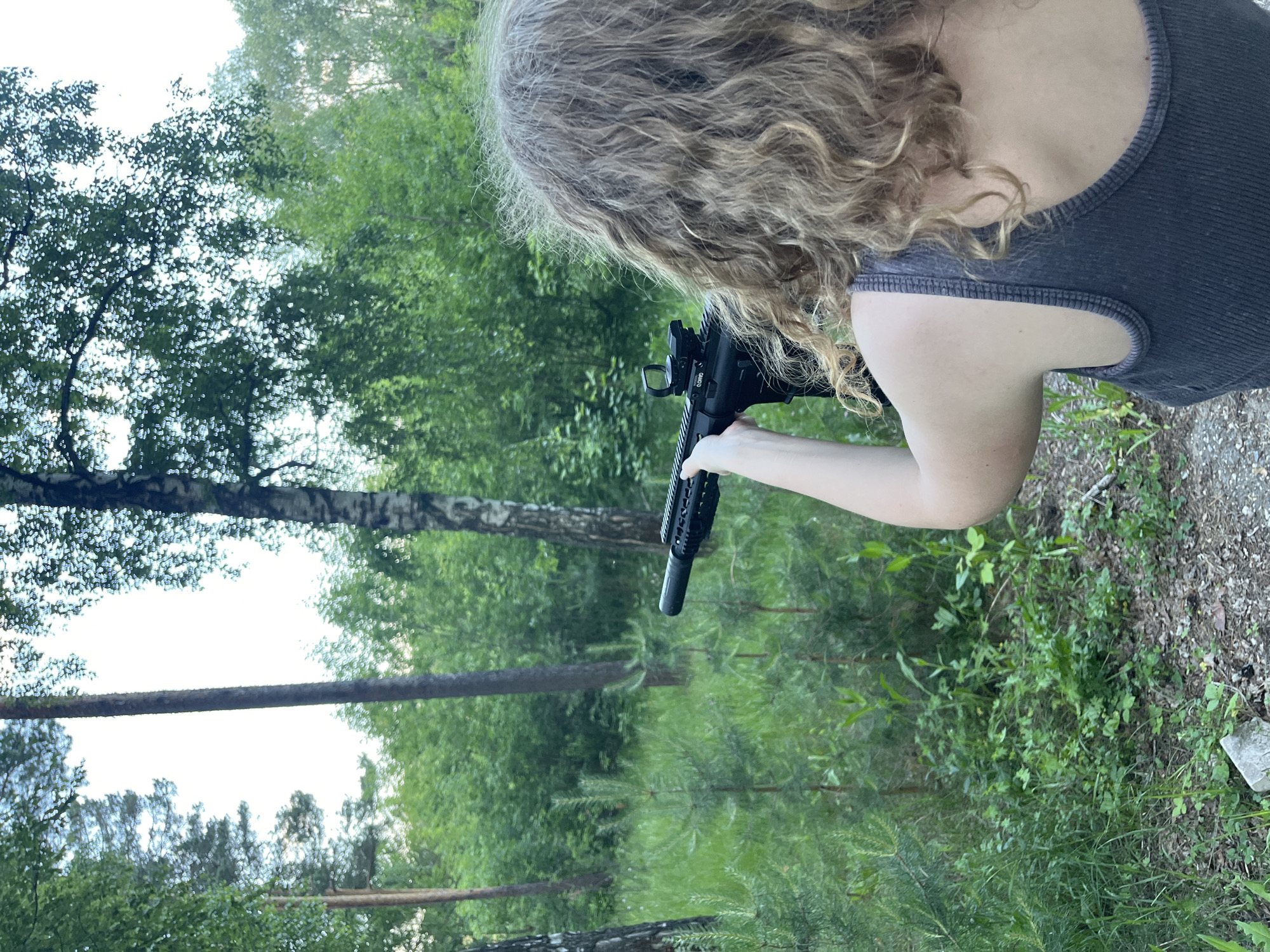 A person is aiming a rifle in a wooded area, with lush green foliage and trees surrounding them. The person is seen from behind, with their right arm extended, holding the rifle. The rifle appears to be a black assault rifle with a scope. The person has curly blonde hair and is wearing a dark grey tank top. The environment is natural, with tall trees and undergrowth. The lighting suggests it is daytime, possibly late afternoon or early morning, given the soft light. There are no other people visible in the image. The ground is covered in dirt, leaves, and some small stones.