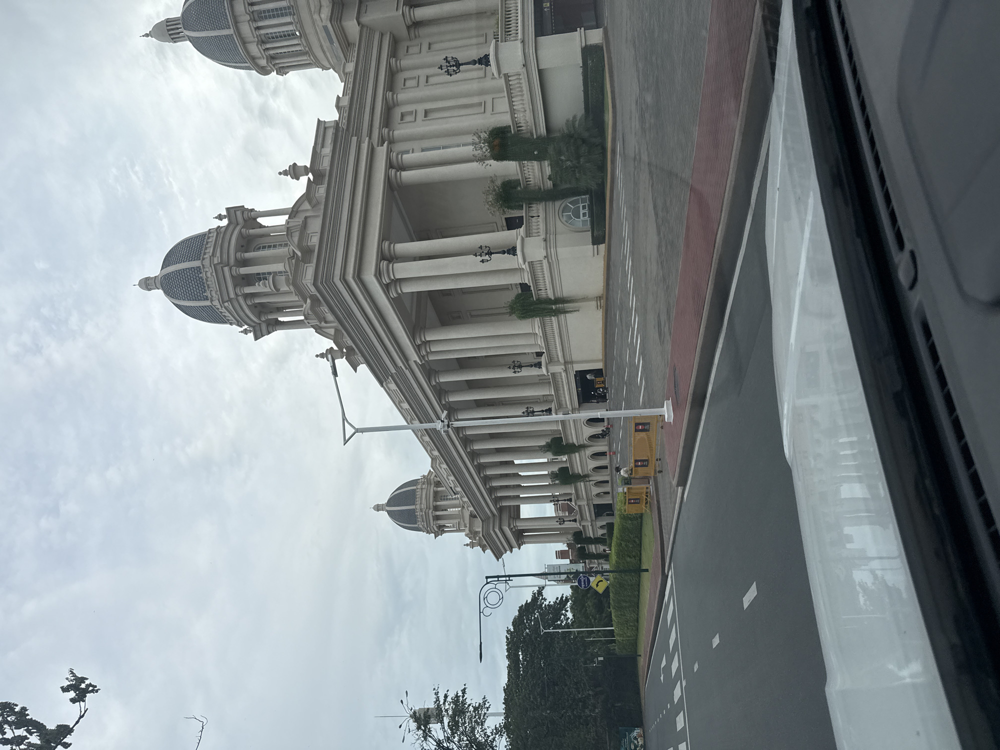 The image shows a wide-angle exterior view of a large, ornate building with classical architecture, including numerous columns and domes, situated in the city of Khan Chbar Ampov, Cambodia. The building is painted in a light cream color and features dark blue domes. In the foreground, a paved road with white lane markings is visible, along with a pedestrian crossing. A white vehicle's dashboard and windshield are in the immediate foreground, suggesting the photo was taken from inside a car. Yellow barriers are placed along the roadside in front of the building. Several people are present near the entrance of the building, and some are visible in the distance walking along the sidewalk. Trees and greenery surround the building and the roadside. The sky is overcast with clouds.