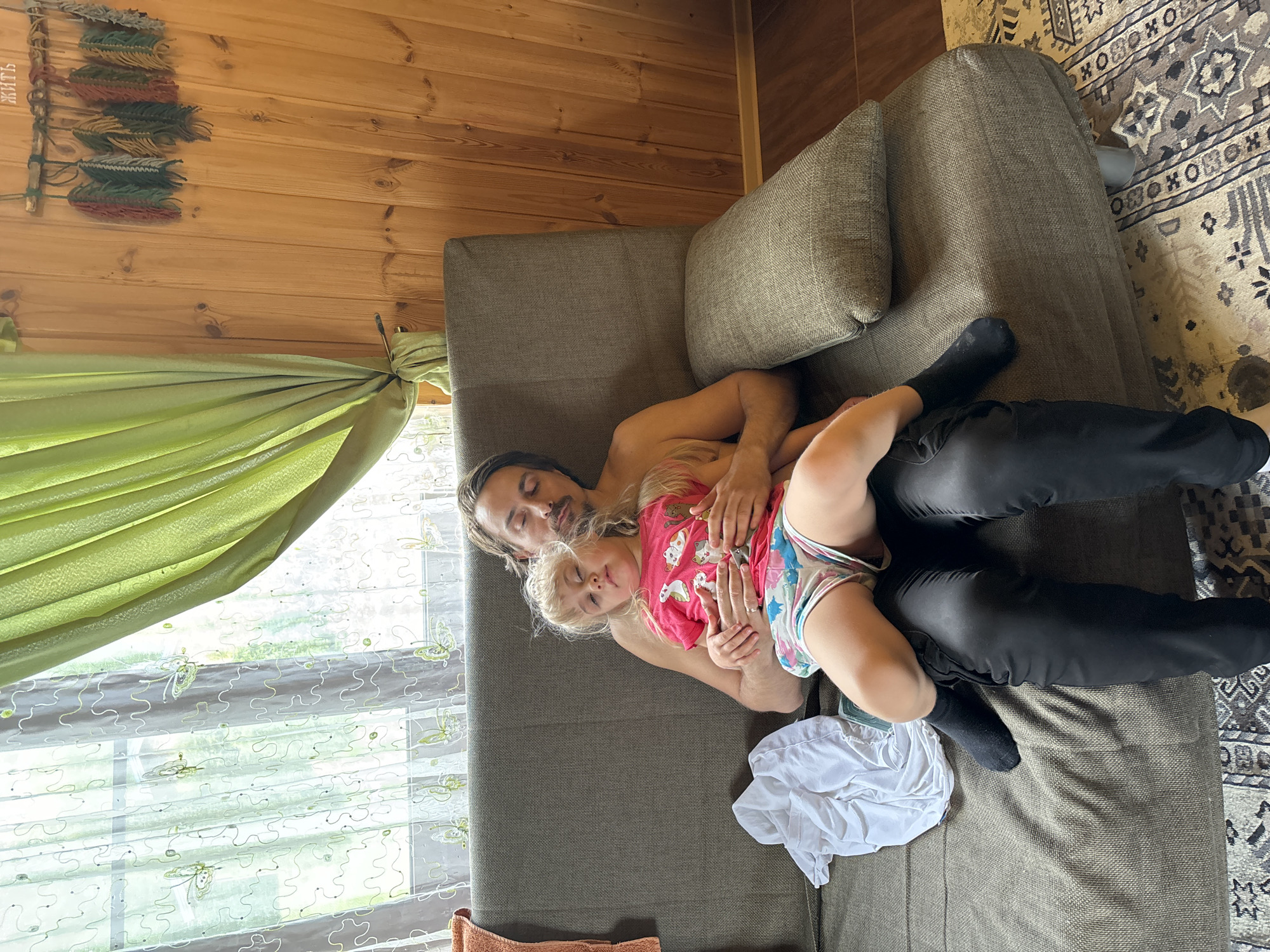 A man and a young child are lying on a gray couch indoors. The man is wearing black pants and socks, but is shirtless. The child is wearing a pink shirt with cat images and patterned shorts. They are positioned near a window covered with a sheer, patterned curtain. To the left of the window is a wooden wall, with a decorative wall hanging. A rug with geometric patterns is visible on the floor next to the couch. The location is Unknown, Unknown.