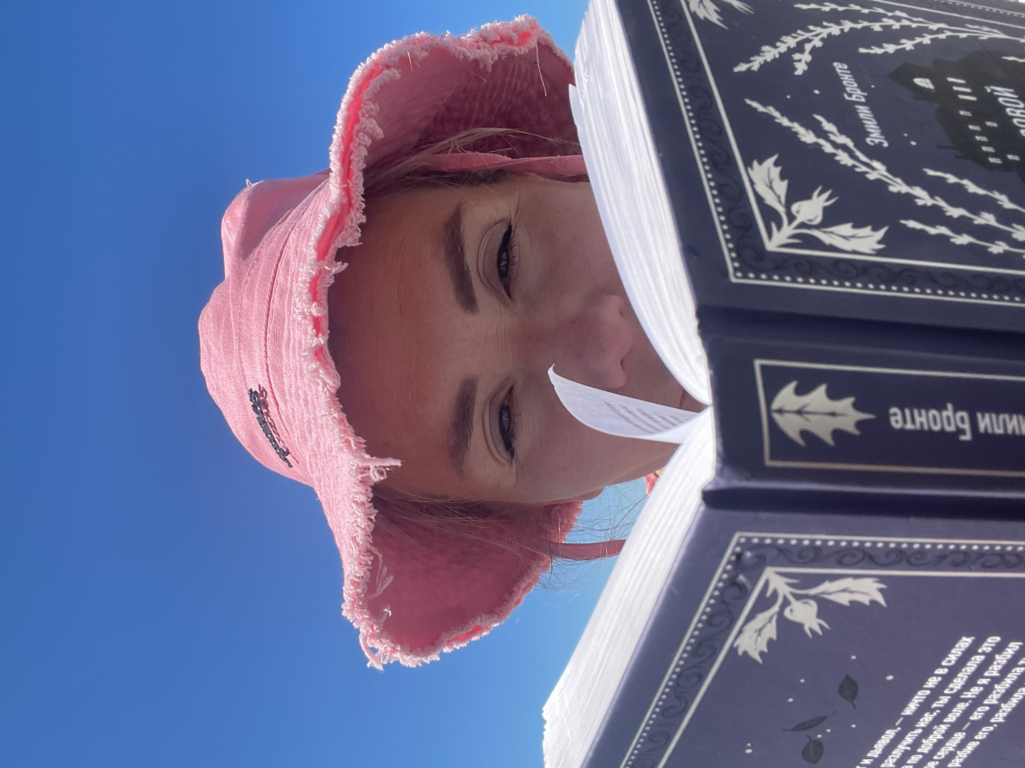 A woman wearing a pink bucket hat reads a book outdoors under a bright blue sky. The woman's face is partially obscured by the book, but her eyes are visible, looking down at the pages. The book is open, revealing text and decorative elements on the pages. The scene suggests a relaxed and leisurely activity, possibly in a park or other outdoor setting.